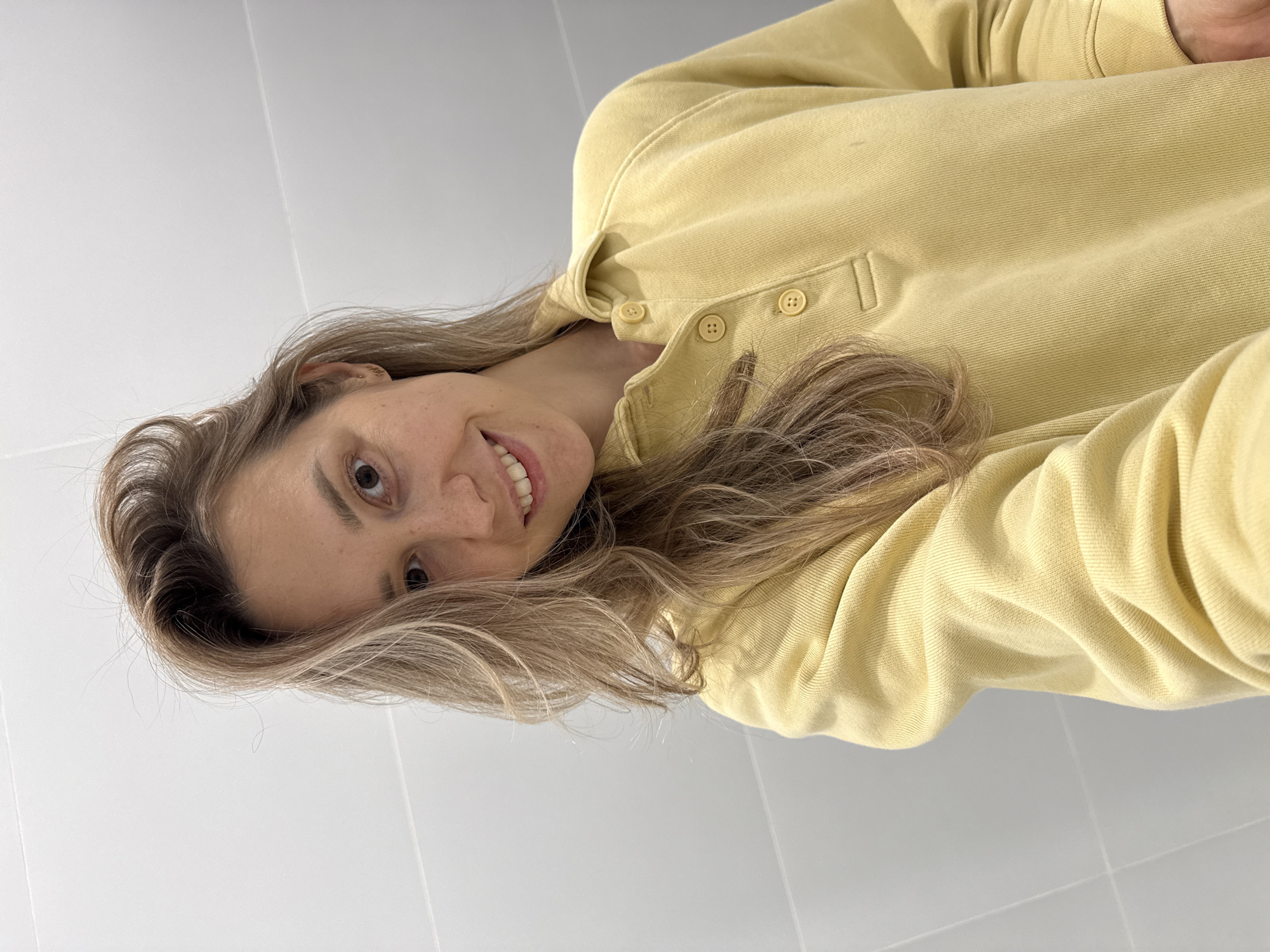 A headshot, likely a selfie, features a person, possibly female, with light blonde to light brown wavy hair. The person is looking directly into the camera with a gentle smile, their head tilted. They are wearing a light yellow long-sleeved shirt or sweatshirt with a collar and a placket featuring three matching yellow buttons. The background is a plain, light-colored surface, possibly a white or light gray wall with subtle grid lines indicating tiling. The lighting is bright and even, suggesting an indoor setting. No specific activity is depicted beyond posing for the photo. The person's expression appears friendly and pleasant. There is no visible text in the image. The location context provided is Новая Казмаска, Russia.