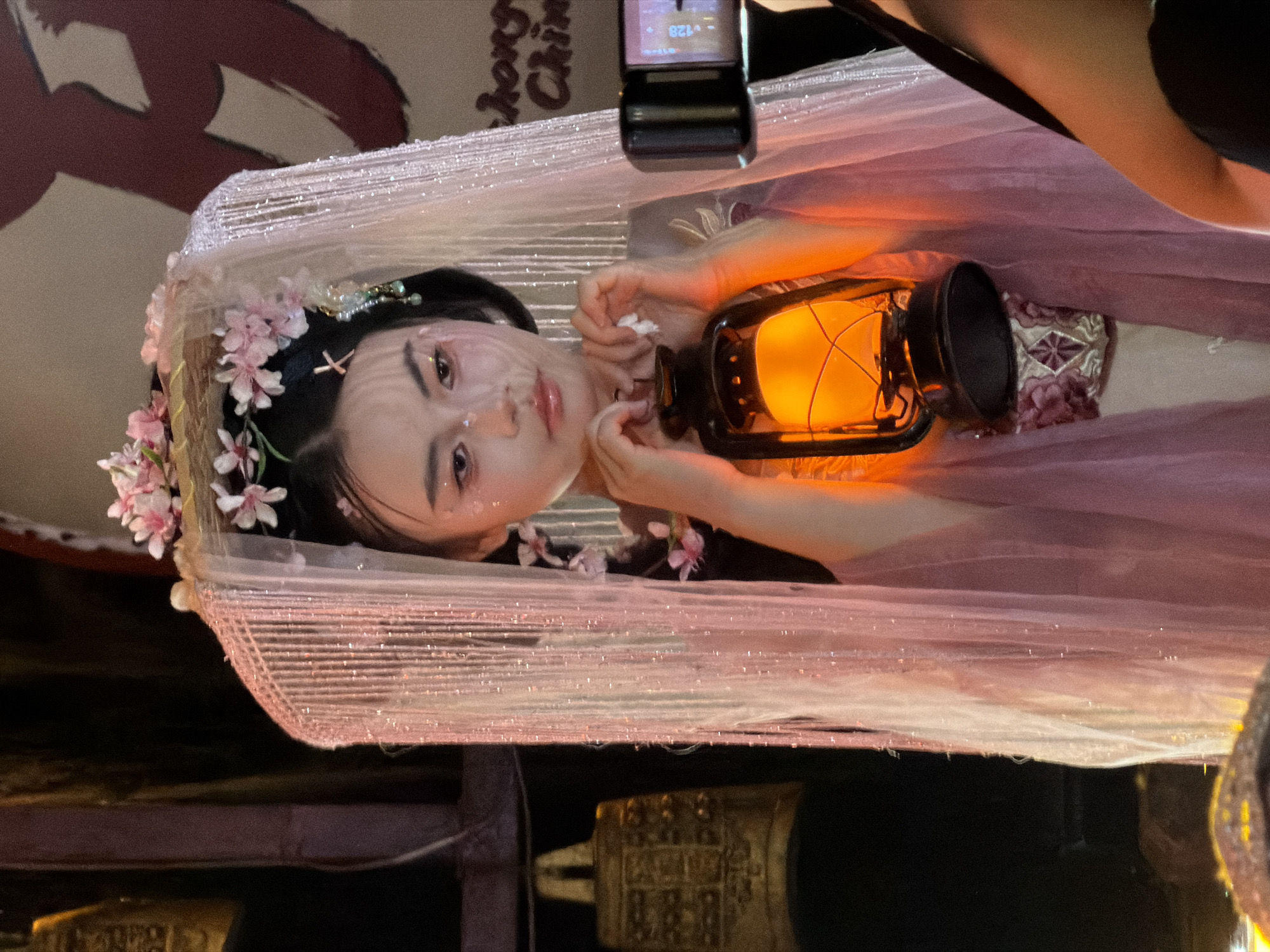 A young East Asian woman in traditional Chinese costume, adorned with pink flowers and a delicate veil, holds an antique oil lantern emitting a warm orange glow. She is positioned in front of a dark background featuring ancient bells and elaborate red Chinese calligraphy. The woman's makeup is dramatic, with glitter accents on her face and pink blush. A camera is visible to the right, suggesting the scene is being filmed or photographed. The location is specified as Yuzhong District, China, implying a cultural or historical setting.