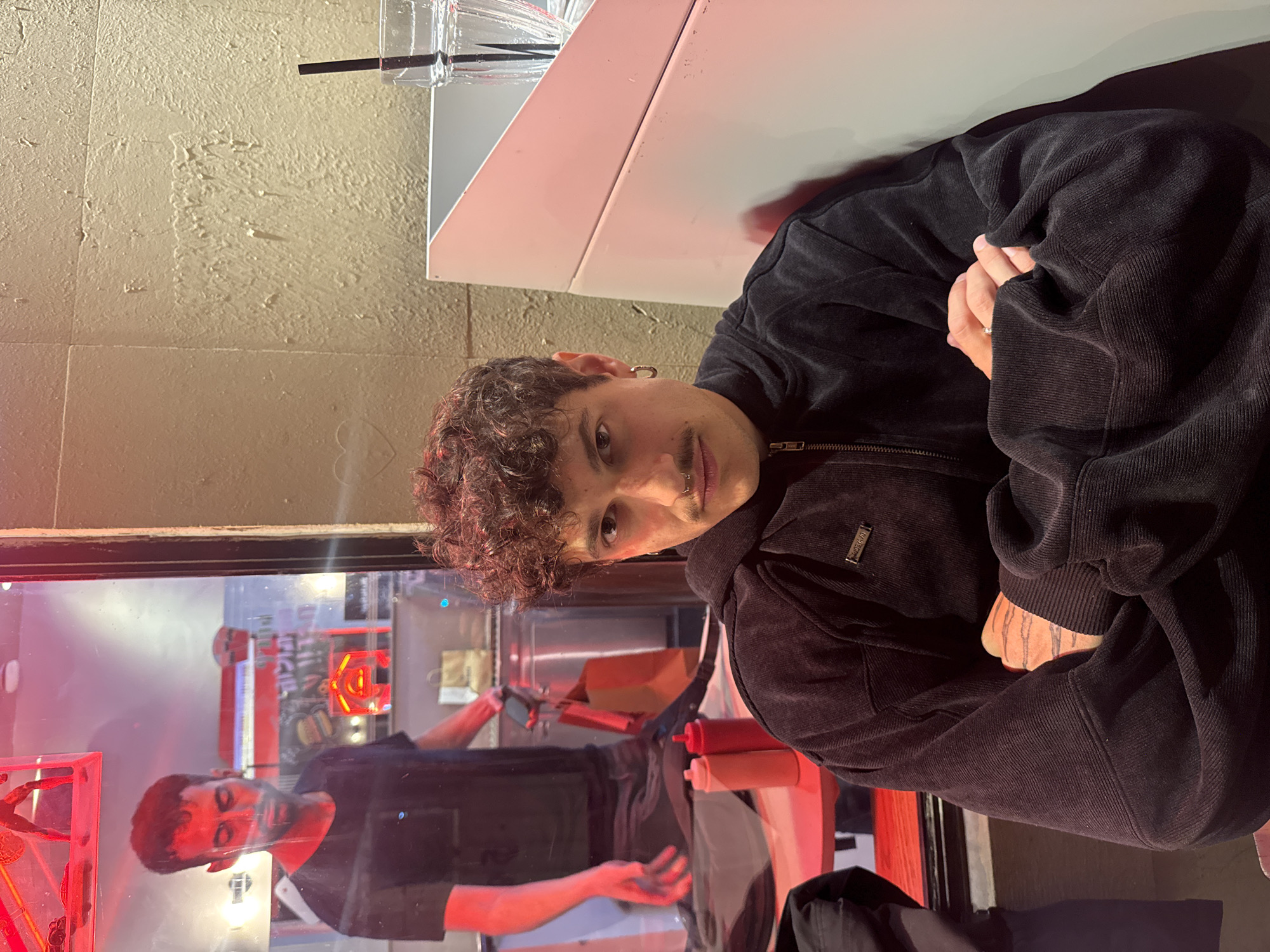 A young person with curly hair and a black corduroy jacket is sitting at a table. They are facing the camera with their hands clasped together. Reflected in a glass behind them, a person wearing a black t-shirt can be seen in what appears to be a restaurant setting with various bottles and red lighting. The location is likely a restaurant or bar in Tel Aviv, Israel, though this cannot be definitively confirmed from the image alone.
