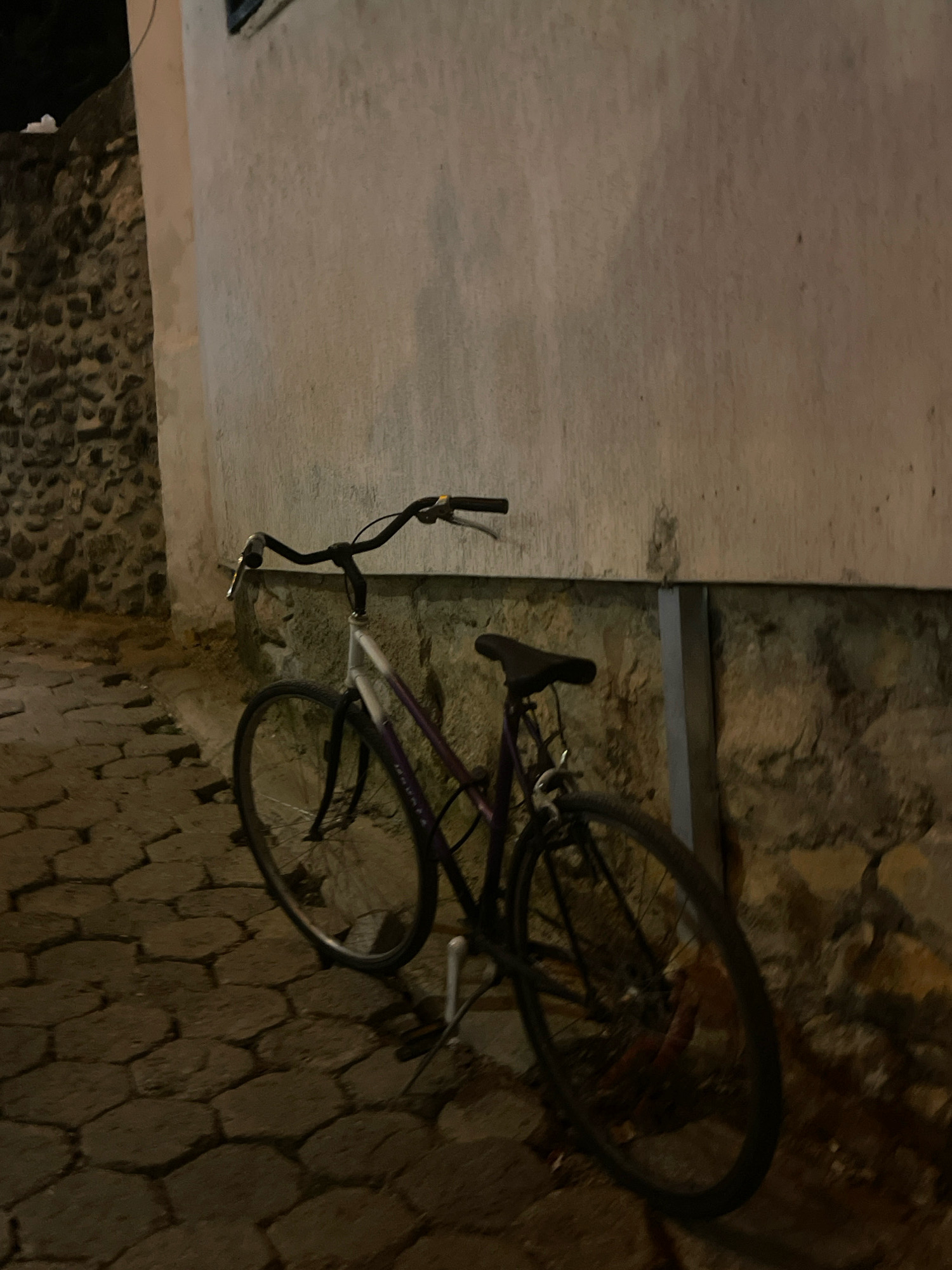 The image depicts a bicycle parked on a cobblestone street in Prizren, Kosovo. The bicycle has a dark frame with purple accents and a white front fork. It is leaning against a textured, light-colored wall that shows signs of weathering. To the left of the bicycle, a stone wall is visible. The lighting suggests it is nighttime or dusk, with shadows cast by the bicycle and the surrounding structures. The street is paved with irregularly shaped cobblestones. No people are present in the scene. The overall atmosphere is quiet and still.