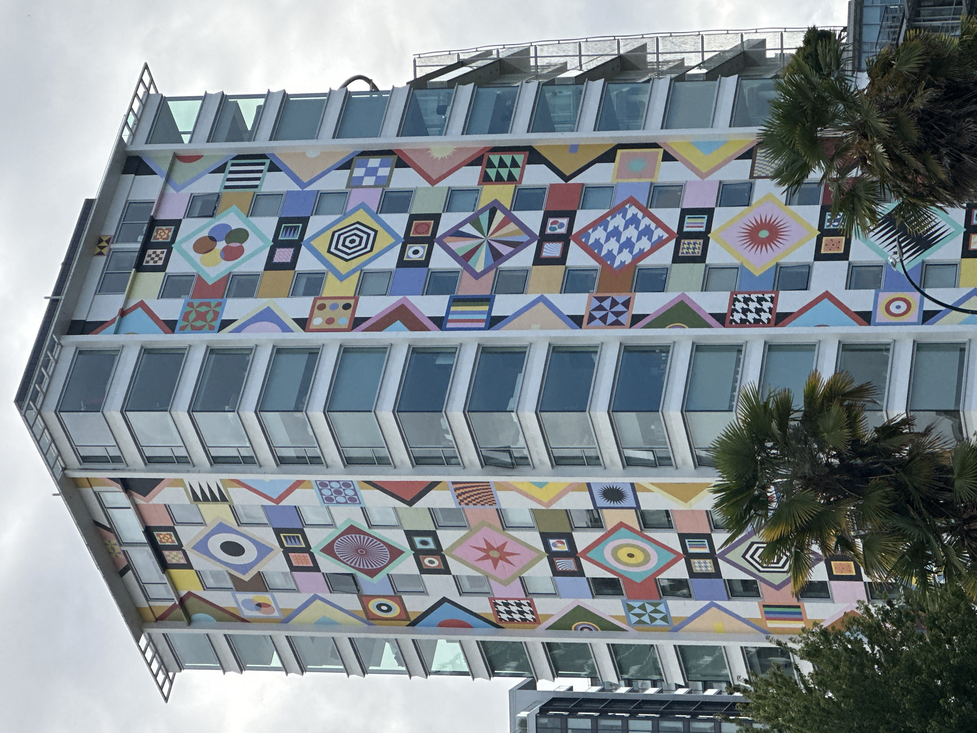 This is a medium-angle, eye-level shot of a brightly decorated, modern apartment building in Vancouver, Canada, with a colorful geometric mural covering a significant portion of its facade. The mural features a vibrant and complex design composed of various shapes and patterns, including diamonds, squares, circles, and stripes, in a multitude of colors such as yellow, pink, blue, green, red, and black. These patterns are organized into distinct blocks, creating a patchwork effect against the building's white exterior. The building itself is a multi-story structure with numerous glass-paneled windows and balconies that reflect the overcast sky. Palm trees and other foliage are visible in the foreground, partially obscuring the lower levels of the building and suggesting a temperate climate. The overall scene is one of urban architecture punctuated by bold, artistic expression.