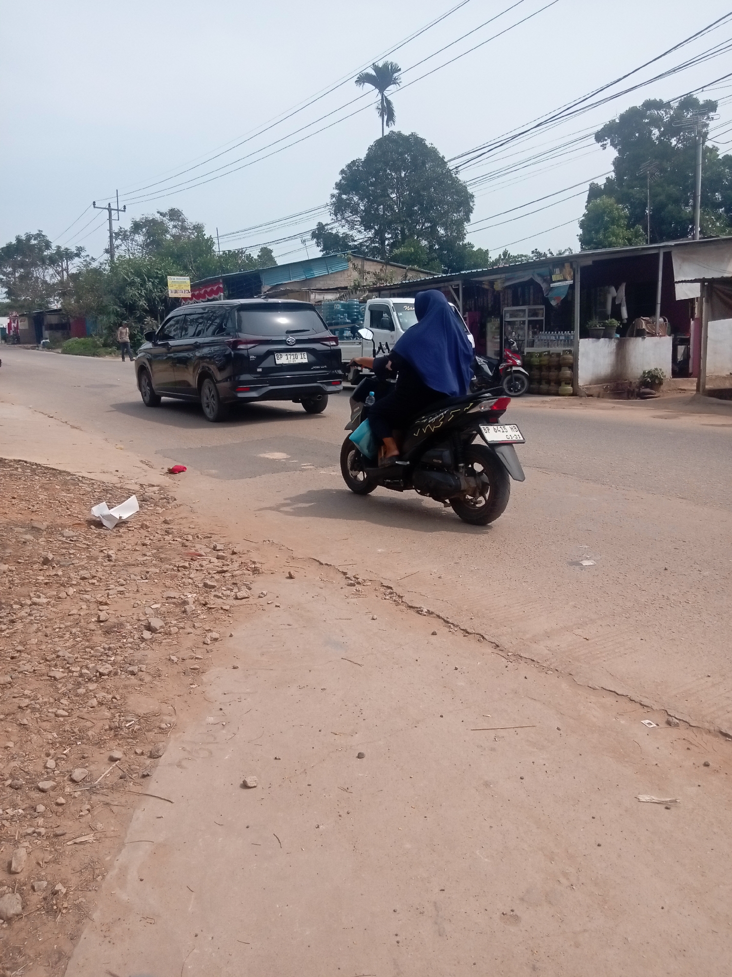 A black SUV and a person on a motorcycle are seen on a paved road in Batam, Indonesia. The SUV, a Daihatsu, has a license plate reading "BP 3739 IE". The motorcycle, a black scooter, has a license plate reading "BP 6415 HB". The person on the motorcycle is wearing a blue hijab and a dark-colored outfit. The road is lined with various shops and vegetation. Some litter is present on the side of the road, including a white crumpled piece of paper and a red object. The sky is overcast.