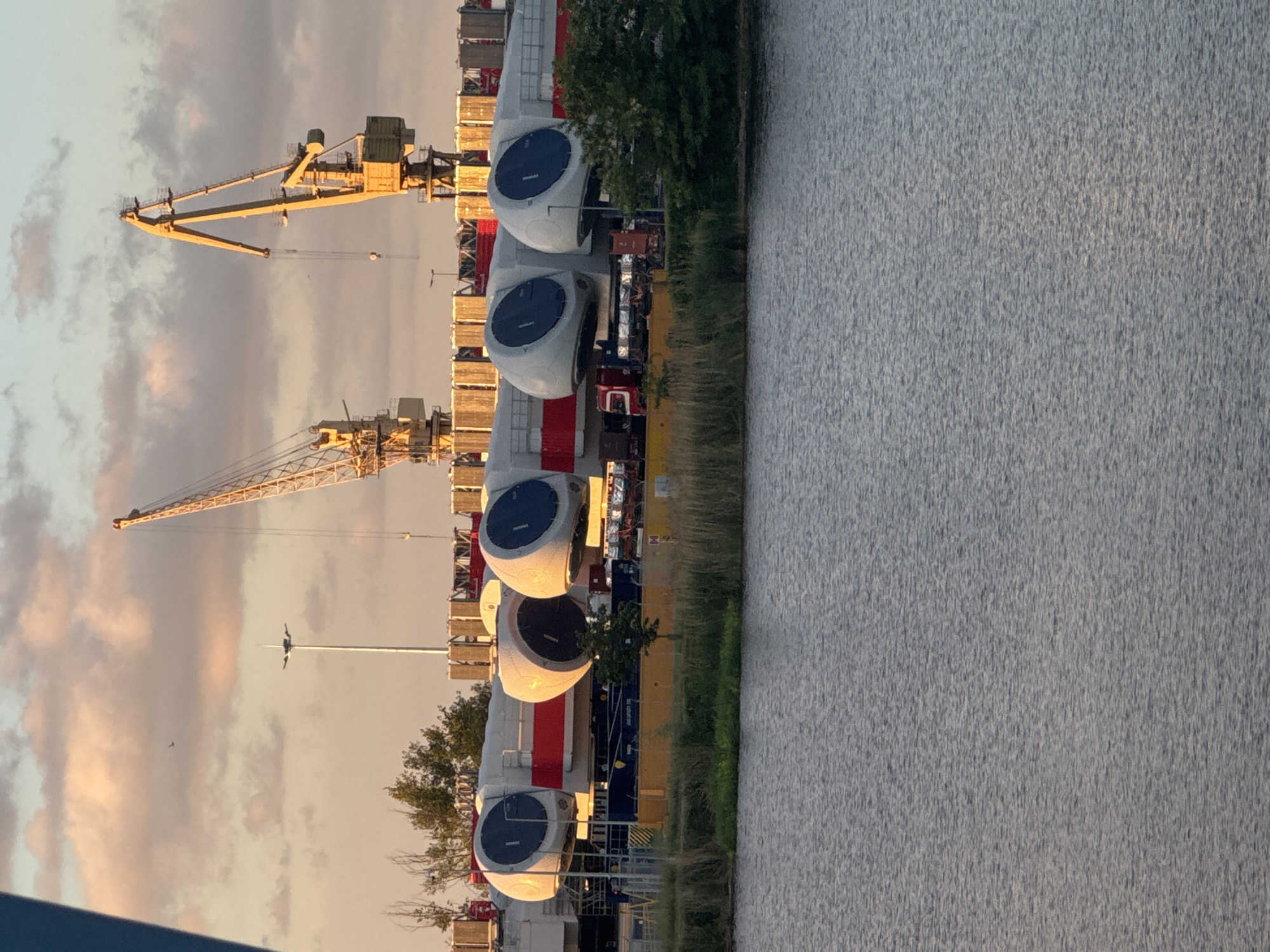 The image shows a row of wind turbine nacelles being transported near a body of water, likely the Oder River, in Szczecin, Poland. The nacelles are cylindrical, white, and red with a dark blue center. The nacelles are on yellow transport carriers. There are two yellow harbor cranes in the background. The sky is cloudy.