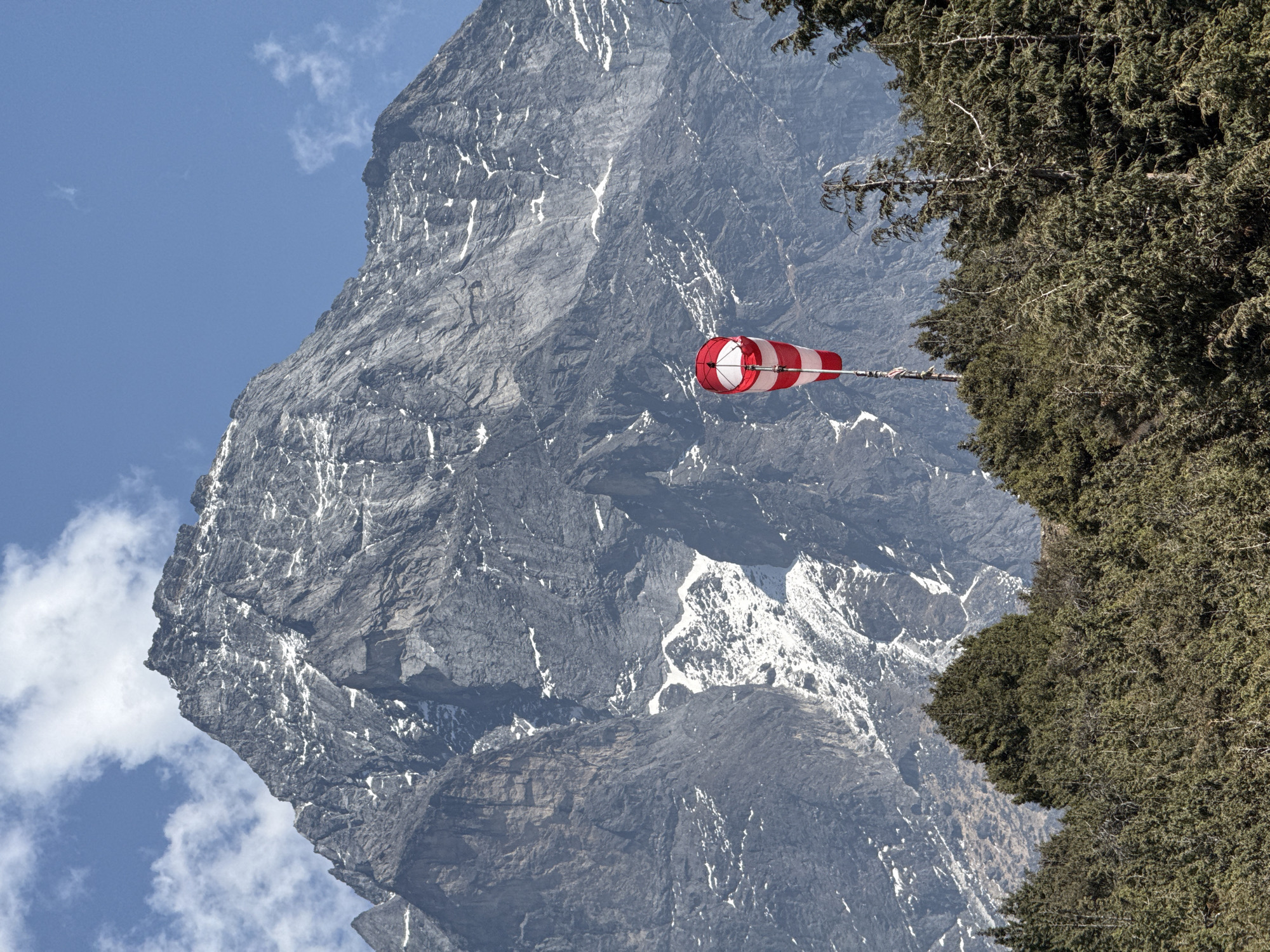 The image captures a landscape near Namche, Nepal. A large, rocky mountain occupies the left side of the frame, displaying prominent ridges and patches of snow. The blue sky is visible beside the peak, with a few clouds. A windsock with alternating red and white stripes is visible near the center. A dense evergreen tree appears on the right side of the frame.