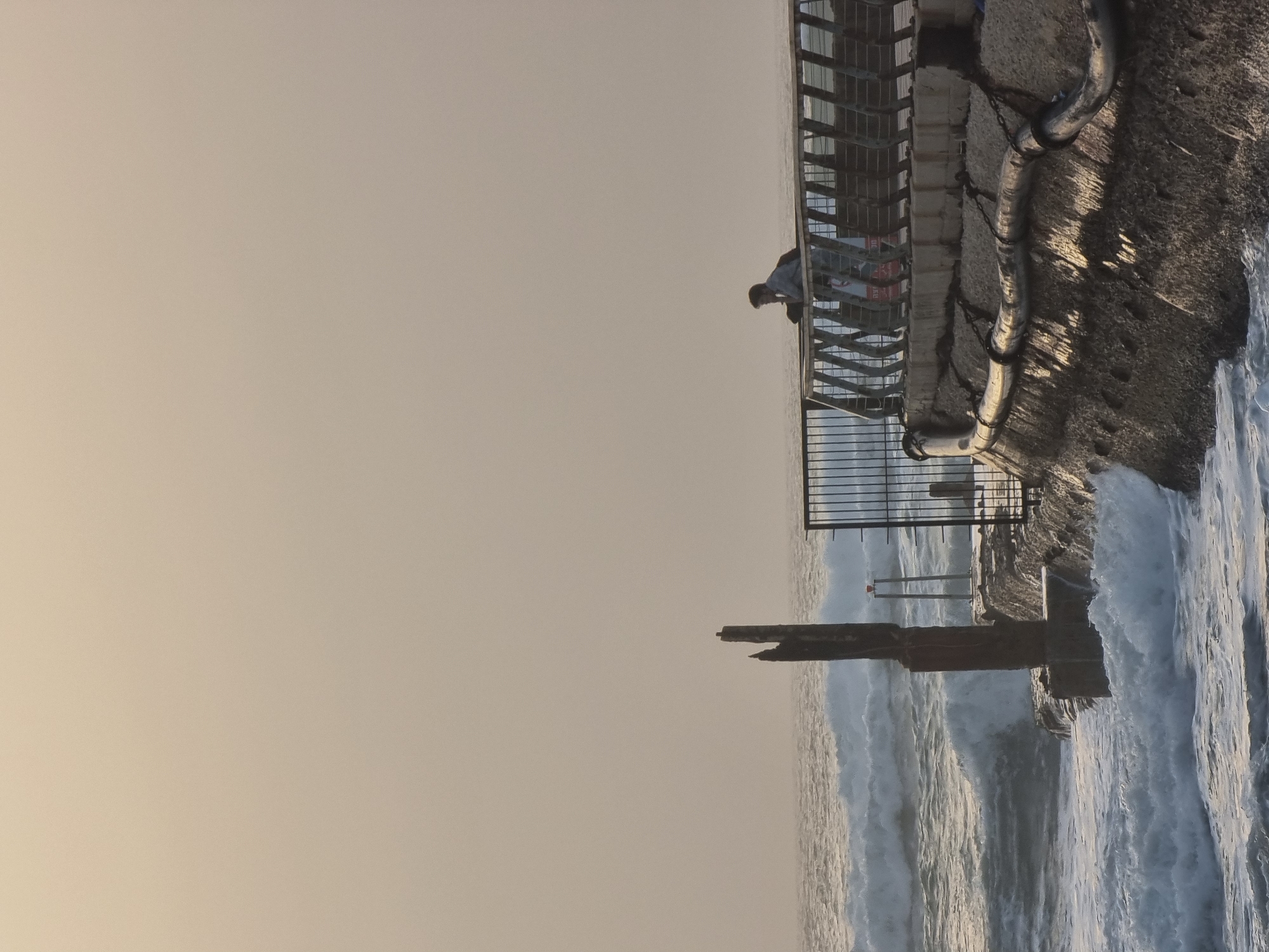 A vertical outdoor shot depicts the seawall at Tel Aviv, Israel, with a person at a railing. The ocean is rough, with large waves crashing against the wall and a partially submerged structure. The sky is overcast, creating soft lighting. The seawall, made of concrete and metal railings, shows signs of wear. The person at the railing appears to be observing the sea. A metal pipe runs along the top of the wall.