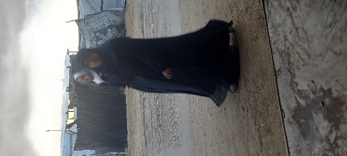 Two women wearing traditional Islamic dress stand next to each other. The woman in front has a white hijab, and the woman behind her is wearing a black abaya. They are outside, possibly in a refugee camp. Tents or temporary shelters can be seen in the background. The ground appears to be sandy or dirt. The sky is bright, suggesting either early morning or late afternoon.