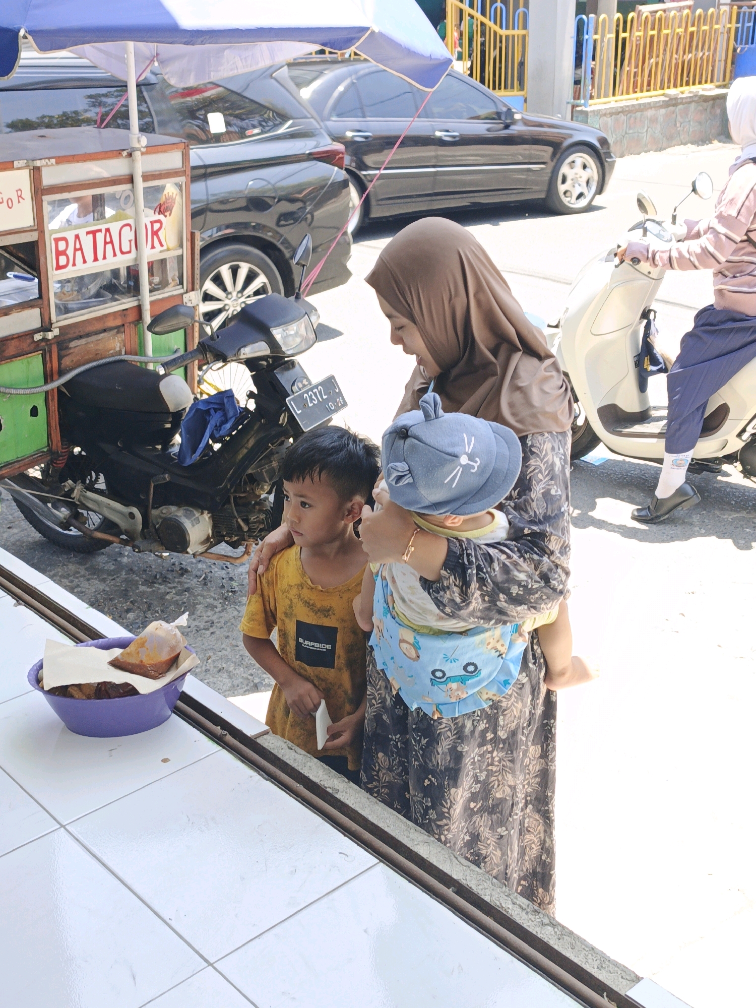 A woman wearing a brown hijab and a floral-patterned dress is holding a baby in a blue carrier. The baby is wearing a blue hat with cat ears and a yellow shirt. To the left of the woman, a young boy in a yellow t-shirt stands and looks to his left. In front of the boy is a purple bowl containing food wrapped in paper, with a plastic bag of sauce on top. Behind them is a food cart with "BATAGOR" written on it in red lettering. A motorcycle is parked next to the food cart, with a license plate visible. In the background, two cars are parked, and a yellow fence with blue poles is present. The scene appears to be outdoors on a sunny day, possibly in the city of Ketegan, Indonesia, indicated by the "BATAGOR" food item, a popular Indonesian street food.