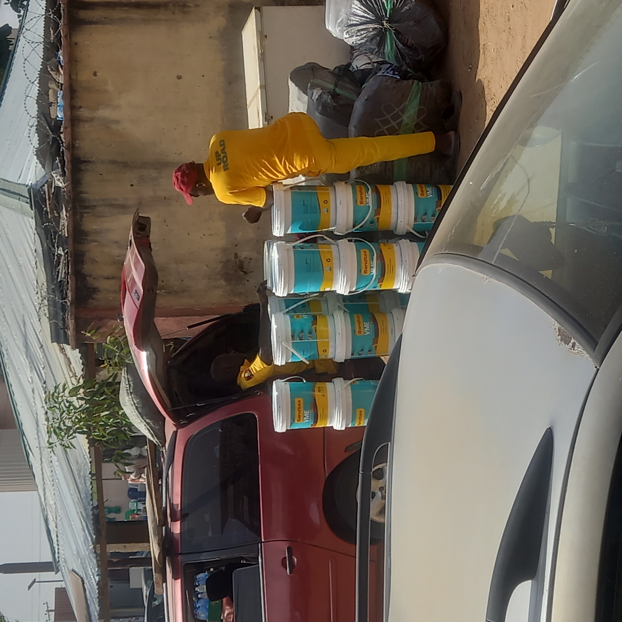 In Dadi Riba, Nigeria, a man wearing a yellow shirt with the words "UP ROAD" on the back and a red cap is loading multiple buckets of "Bandotex VME" paint into the trunk of a red vehicle. Bags of items covered in plastic are next to the paint buckets. A white vehicle is partially visible to the right. A building and barbed wire fence are in the background.
