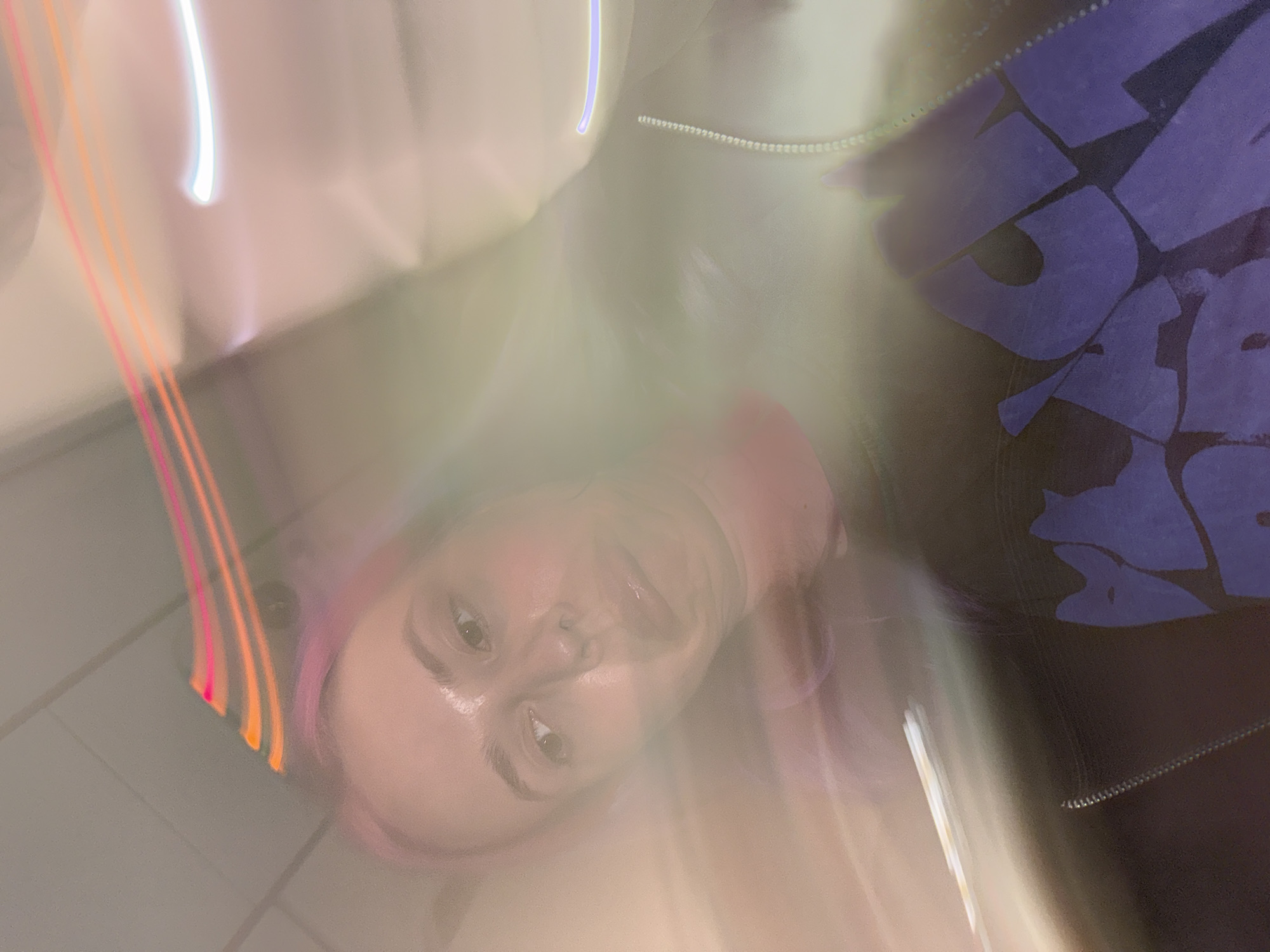 The image is an out-of-focus selfie of a person with pink hair, wearing a dark jacket with large, light-colored lettering, likely taken in City of Zagreb, Croatia. The person is looking directly at the camera with a slight smile. The lighting is uneven and blurry, with streaks of light in the background. The background features a gray wall or surface.
