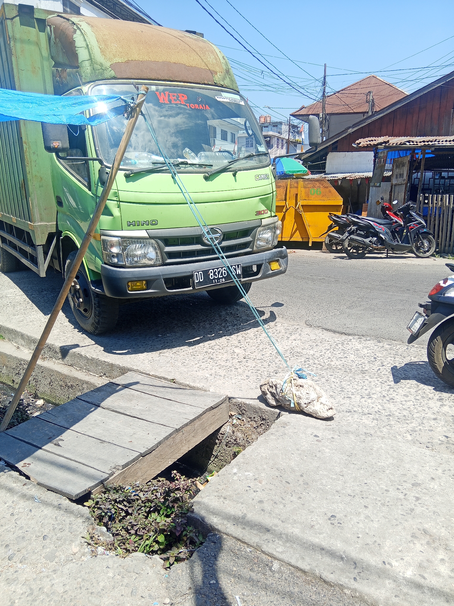 In Makale, Indonesia, on a sunny day, a light green HINO truck is parked on a street next to a sidewalk. A blue tarp is draped over the front of the truck, supported by a wooden pole. A large rock, tied with rope and yellow plastic straps, is placed on the sidewalk and connected by a light blue rope to the truck's grille. Two motorcycles are parked on the street behind the truck, and a yellow dumpster is visible further back. Buildings with reddish-brown roofs line the street, and power lines crisscross the sky. The sidewalk has a small wooden bridge over a ditch filled with plants and debris.