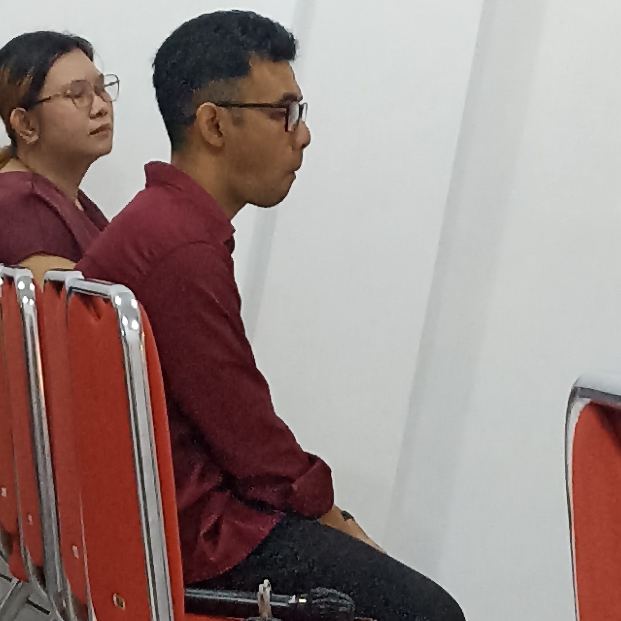 Two individuals are seated in a room with white walls and red chairs with chrome frames. The person on the left, partially visible, is a woman wearing glasses and a maroon shirt. The person on the right is a man with dark, short hair and glasses, also wearing a maroon shirt and black pants. He is looking to his right. The scene appears to be indoors, possibly in a meeting room or waiting area. The lighting is bright. The image quality is moderate, with some pixelation. The location in Magelang, Indonesia, is not specified within the image itself.