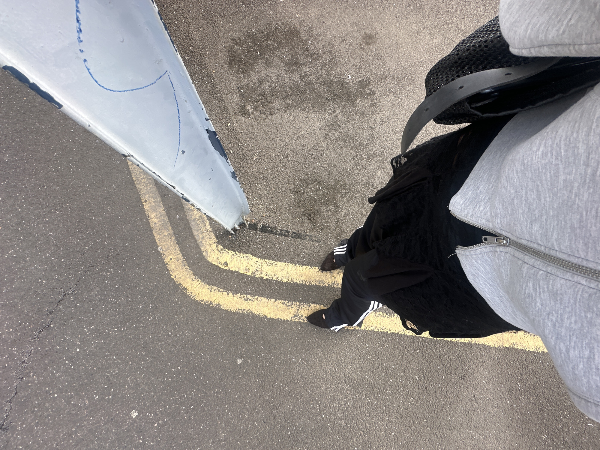A first-person perspective photograph shows the lower body of a person standing on asphalt marked with double yellow lines. The person is wearing black Adidas track pants with white stripes, black footwear, a black mesh bag with a black leather belt over it, and a gray zip-up hoodie. To the left of the person, there is a white metal structure with blue markings on it. The asphalt surface has patches of discoloration. The photograph was likely taken outdoors. Without additional information, the specific location in Tunbridge Wells, United Kingdom, cannot be definitively determined.