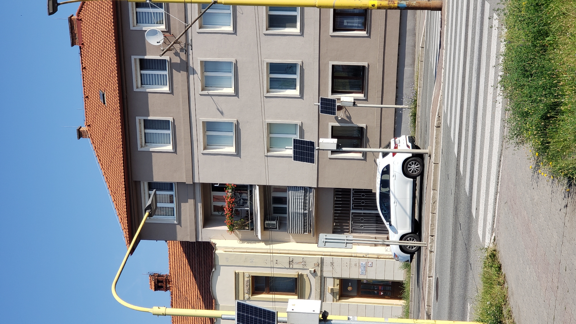 A white SUV is parked at a pedestrian crossing in front of a multi-story building with orange tiled roofs and beige and gray facades. The building has multiple windows, some with blinds, and balconies. There are solar panels mounted on poles in front of the building. A yellow lamppost with a curved arm arches over the street. The sky is clear and blue. The scene appears to be in a city, likely Prešov, Slovakia.