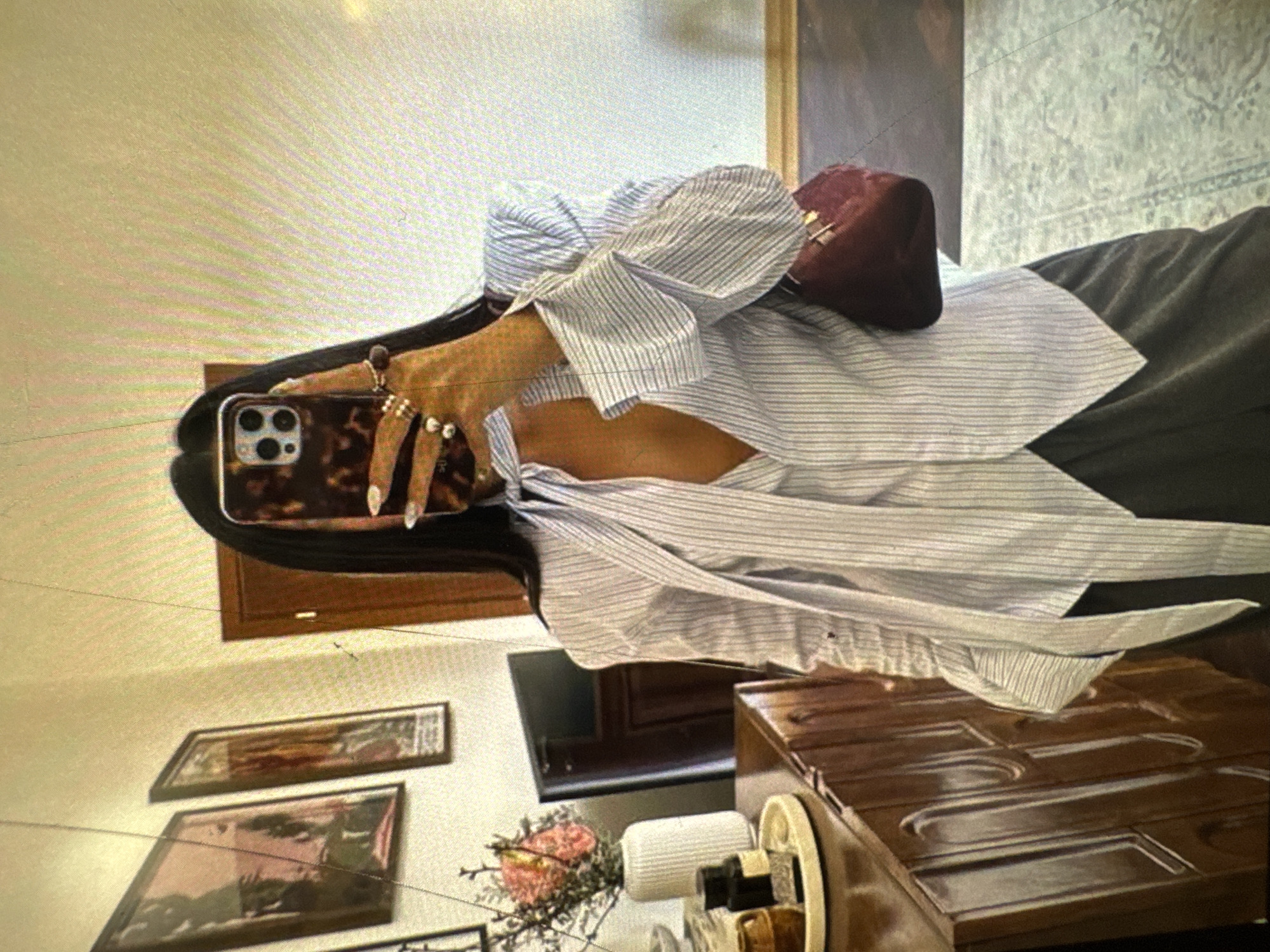 A person is taking a selfie in a dimly lit room. The person is holding up a smartphone with a tortoiseshell case to take the photo. The person is wearing a white and blue striped button-down shirt with a large bow at the neck and a dark-colored skirt. The background shows a wooden dresser, a vase of flowers, and framed pictures on the wall. The floor appears to be tiled.