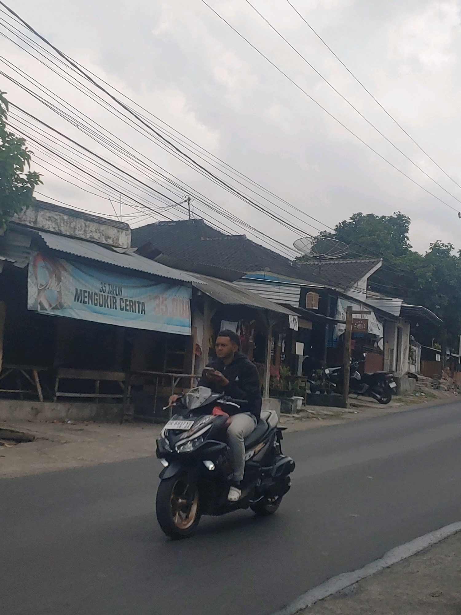 A man rides a dark-colored scooter on a paved road in Kembang Kerang, Indonesia. The man is wearing a dark hoodie and light-colored pants. He is looking down at his phone, which he holds in his left hand. The scooter has a license plate that reads "DK 6937 YV". In the background, there are several buildings with corrugated metal roofs. A banner hangs on the front of one of the buildings, displaying text in Indonesian. The sky is overcast.