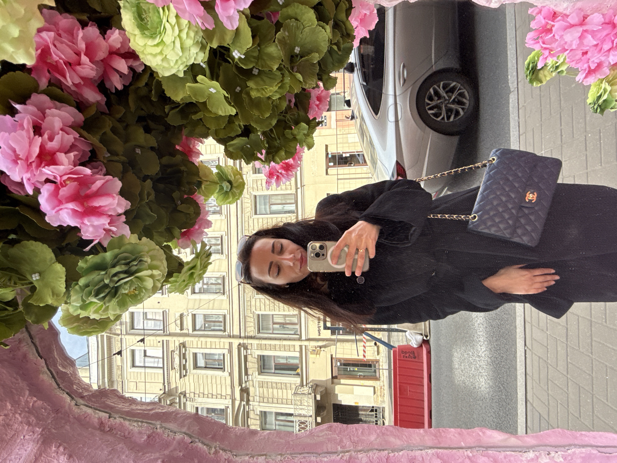 A woman is taking a selfie in a mirror decorated with pink and green flowers. She is wearing a black coat and carrying a blue quilted purse with a gold chain. The reflection shows a street scene with a light-colored building with many windows, a silver car, and a red object. The image was taken in Saint Petersburg, Russia.