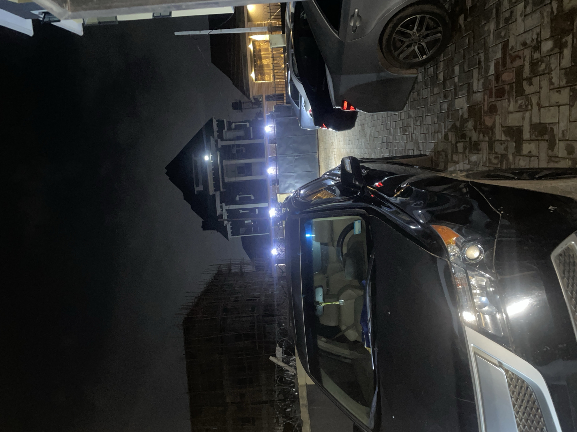 The image captures an outdoor night scene in Abuja, Nigeria, likely a residential or commercial area. In the foreground, the front portion of a black SUV is visible, showcasing its headlights, grille, and windshield. The interior of the SUV, including the beige leather seats, is also discernible. To the right of the SUV, a silver sedan is parked, with its rear visible. Further in the background, there are multiple buildings. One prominent structure appears to be a large, dark house with a distinctive roofline and illuminated windows, suggesting it's a residence. Adjacent to this is another building with a lighter facade and lit windows. To the left, a multi-story building under construction is visible, shrouded in scaffolding. The ground is paved with interlocking brick pavers, some of which appear wet, reflecting the ambient light. The sky above is dark and overcast, typical of nighttime conditions in the region. The scene appears to be a still moment, with no discernible activity or people present in the frame.