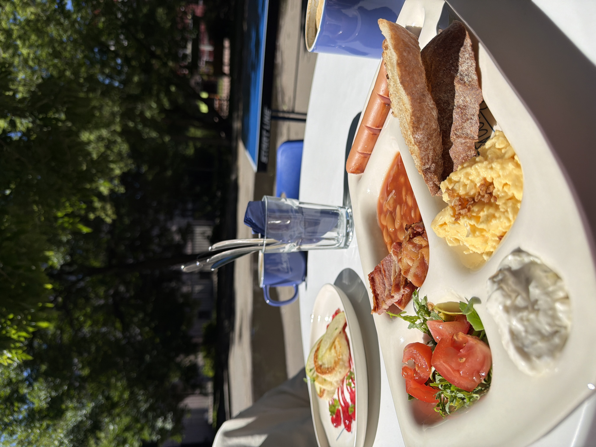 An outdoor cafe scene in Saint Petersburg, Russia, features a bright, sunny day with lush green trees providing dappled shade. The focus is on a breakfast spread on a white table.
On the left, a white plate holds three round, golden-brown pancakes or fritters, topped with thin slices of green apple and drizzled with a vibrant pink sauce.
In the foreground, a large white compartmentalized dish contains a hearty breakfast. The compartments hold:
- Fresh, chopped tomatoes mixed with green herbs.
- A dollop of white cream cheese or sour cream.
- Savory scrambled eggs sprinkled with small brown bits, possibly crispy onions or herbs.
- Several slices of crispy bacon.
- A serving of baked beans in tomato sauce.
- A whole, lightly toasted baguette.
- A single, cooked sausage.
To the right, a blue mug contains coffee or another beverage. A clear glass with cutlery rests behind the food. Another blue mug is visible in the background. The setting appears to be a relaxed outdoor dining area, possibly a cafe patio, with blurred figures and furniture visible in the distance. The overall impression is one of a pleasant, leisurely meal enjoyed al fresco.