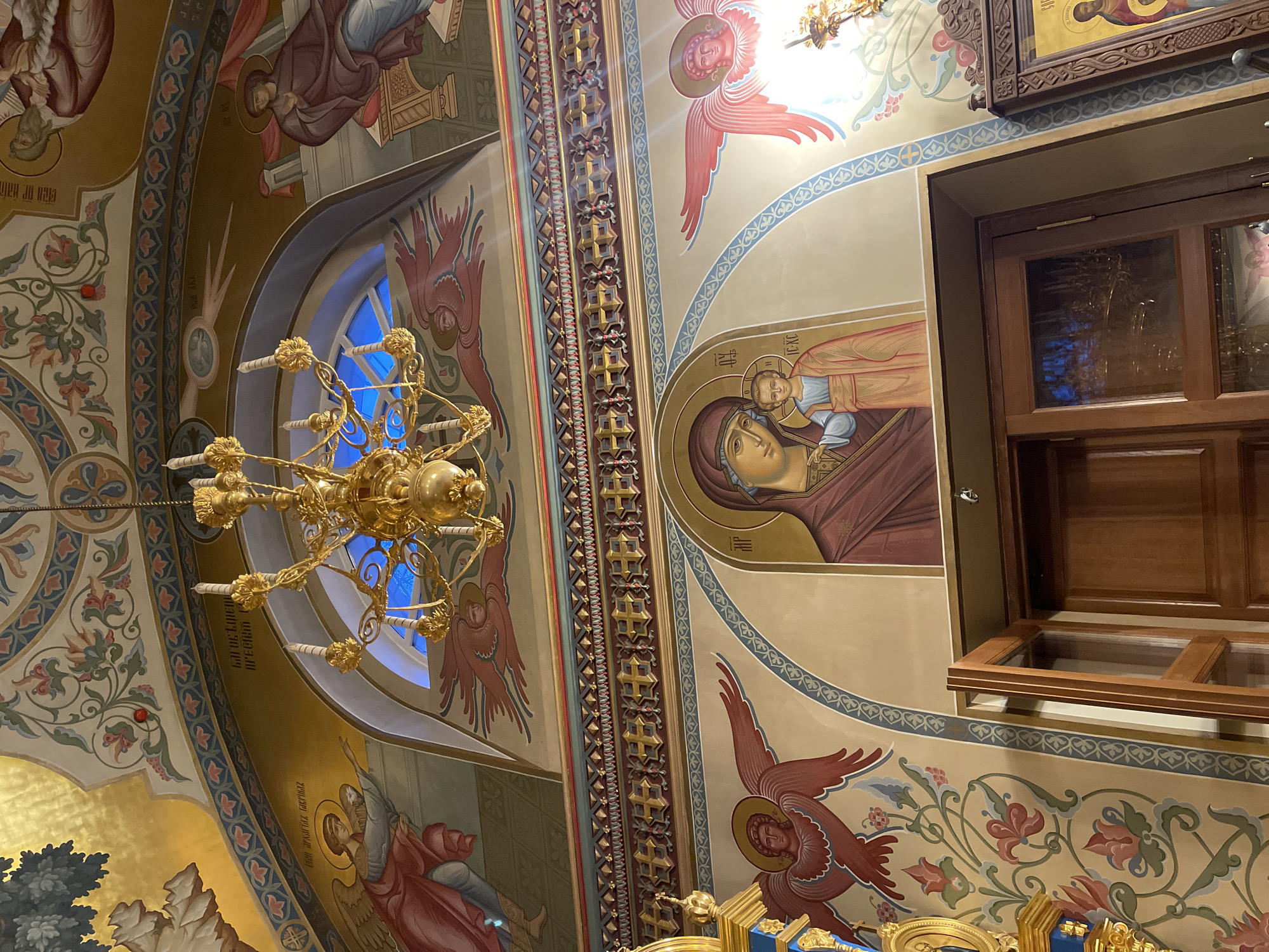 The image shows an interior view of a Russian Orthodox church, potentially located in Moscow, Russia, based on the iconography and architectural style.
The scene is focused on the ornate ceiling, adorned with religious frescoes, including depictions of angels and saints. One fresco shows a Madonna and Child. A gold chandelier hangs from the ceiling.
To the right is a window with a dark wooden frame, reflecting the church's interior.
The walls are decorated with floral motifs and geometric patterns. The overall impression is of a richly decorated and religiously significant space.