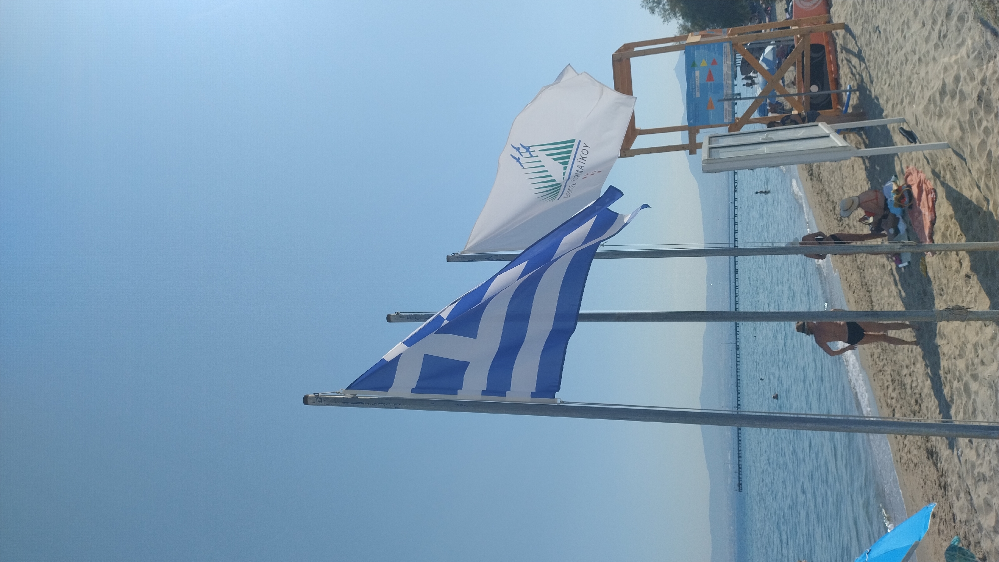The image depicts a beach scene in Perea, Greece, under a clear blue sky. In the foreground, two flags are prominent: the Greek flag, with its blue and white stripes and white cross on a blue canton, flies from a metal pole. To its right, another white flag displays a green design with stylized birds and text.
Behind the flags, a sandy beach stretches towards the blue sea. Several people are present on the beach. Two women are visible in the mid-ground, one in black swimwear facing away from the camera and another in a bikini. Further down the beach, another person is seated, wearing a hat. In the sea, a few individuals can be seen swimming or wading.
In the background, a long pier extends into the water, with a distant shoreline and hills visible. To the right of the flags, a wooden lifeguard tower with a white sign is situated on the sand. The overall scene suggests a sunny day at a coastal resort.