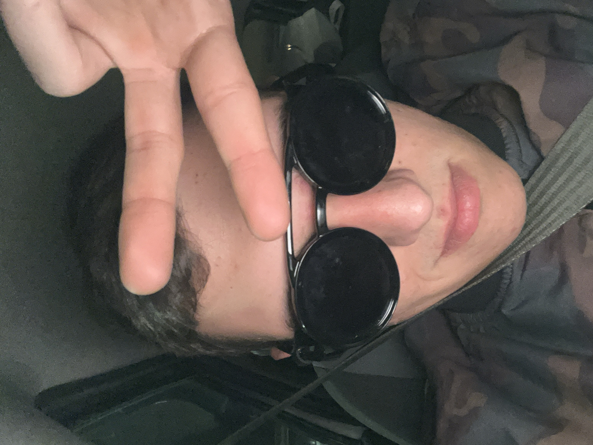 A person is shown from a low, close-up angle in a horizontally flipped image. The person is wearing round, black sunglasses and holding up their hand with two fingers raised in a peace sign, obscuring part of their face. They appear to be inside a vehicle, secured with a seatbelt. The person has short, dark hair and fair skin. Their clothing has a camouflage pattern. The background is indistinct, suggesting the interior of a car. The location is not identifiable from the image.