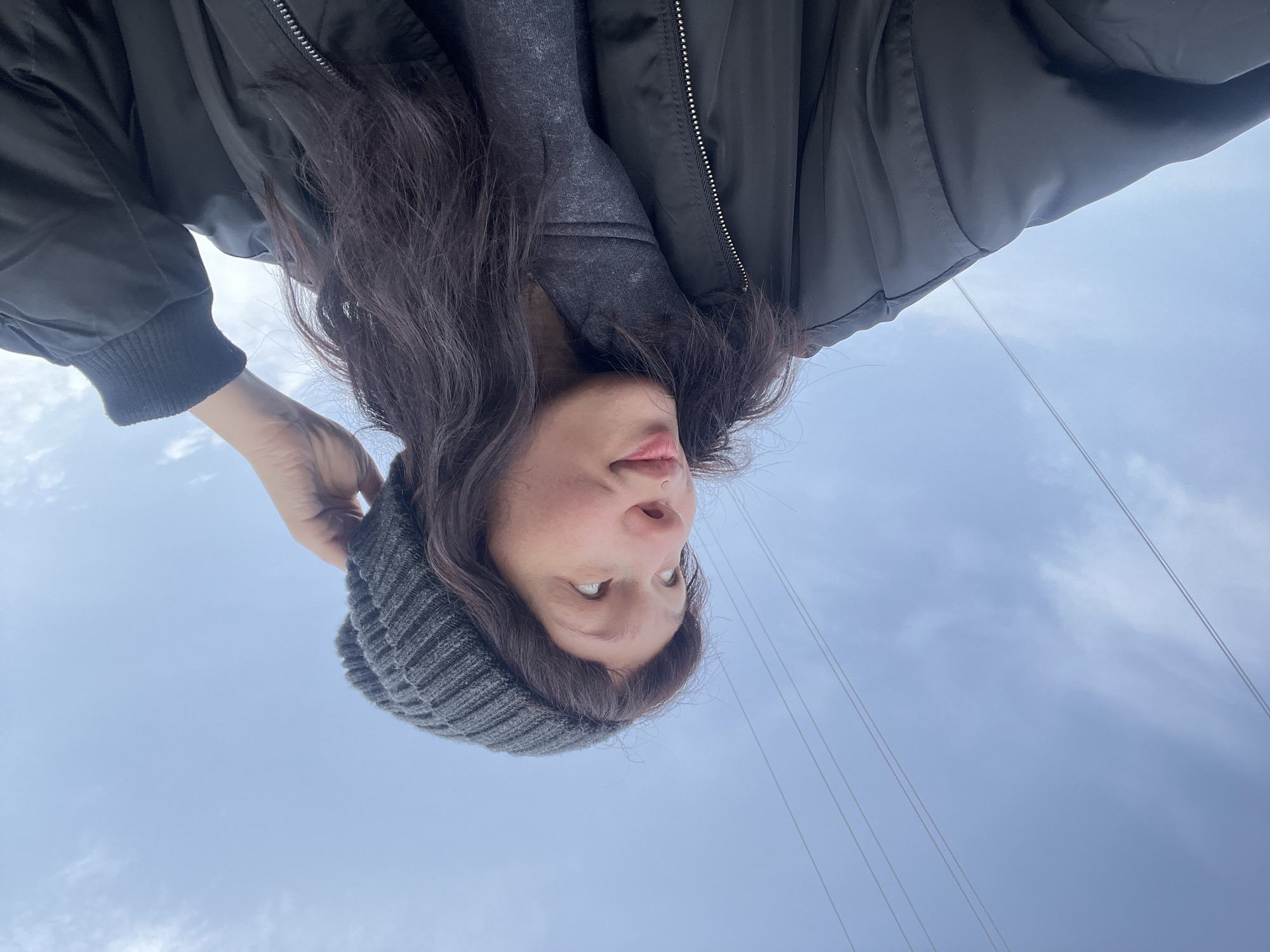 An upward-angled outdoor portrait features a person, likely female, positioned upside down in the frame against a clear to partly cloudy blue sky. The subject has long, dark, wavy hair, and is wearing a dark grey ribbed beanie, a grey scarf or neck warmer, and a black bomber-style jacket with a zippered front. Their face is visible, looking directly upwards (towards the top of the frame), with an open mouth and slightly raised eyebrows, suggesting a curious or playful expression. A subtle nose piercing is also visible. One hand, likely the left, is partially visible near the top of their head, possibly adjusting the beanie or hair. In the background, thin wires, possibly power or communication lines, stretch diagonally across the sky, particularly noticeable on the right side and in the lower left corner of the frame. The sky is bright with soft white clouds, indicating daytime. The scene appears to be set outdoors in a location like Zhigalki, Russia. There is no visible text in the image.