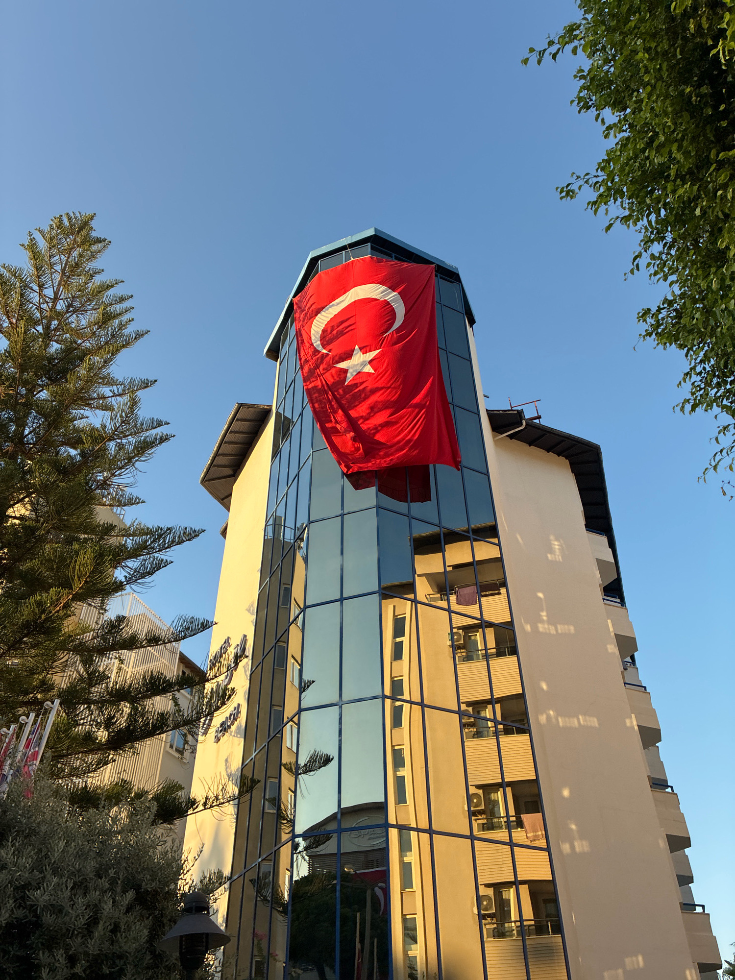 The image shows a large Turkish flag hanging from the top of a modern building with a glass facade in Alanya, Turkey. The building is tall and appears to be a hotel or apartment complex, with balconies visible on its side. The flag is predominantly red with a white star and crescent moon. The sky is clear and blue, suggesting a sunny day. Trees are visible on the left and right sides of the frame, partially obscuring the building. The reflection of the building and the sky can be seen in the glass facade. The overall impression is one of a patriotic display in a coastal city.