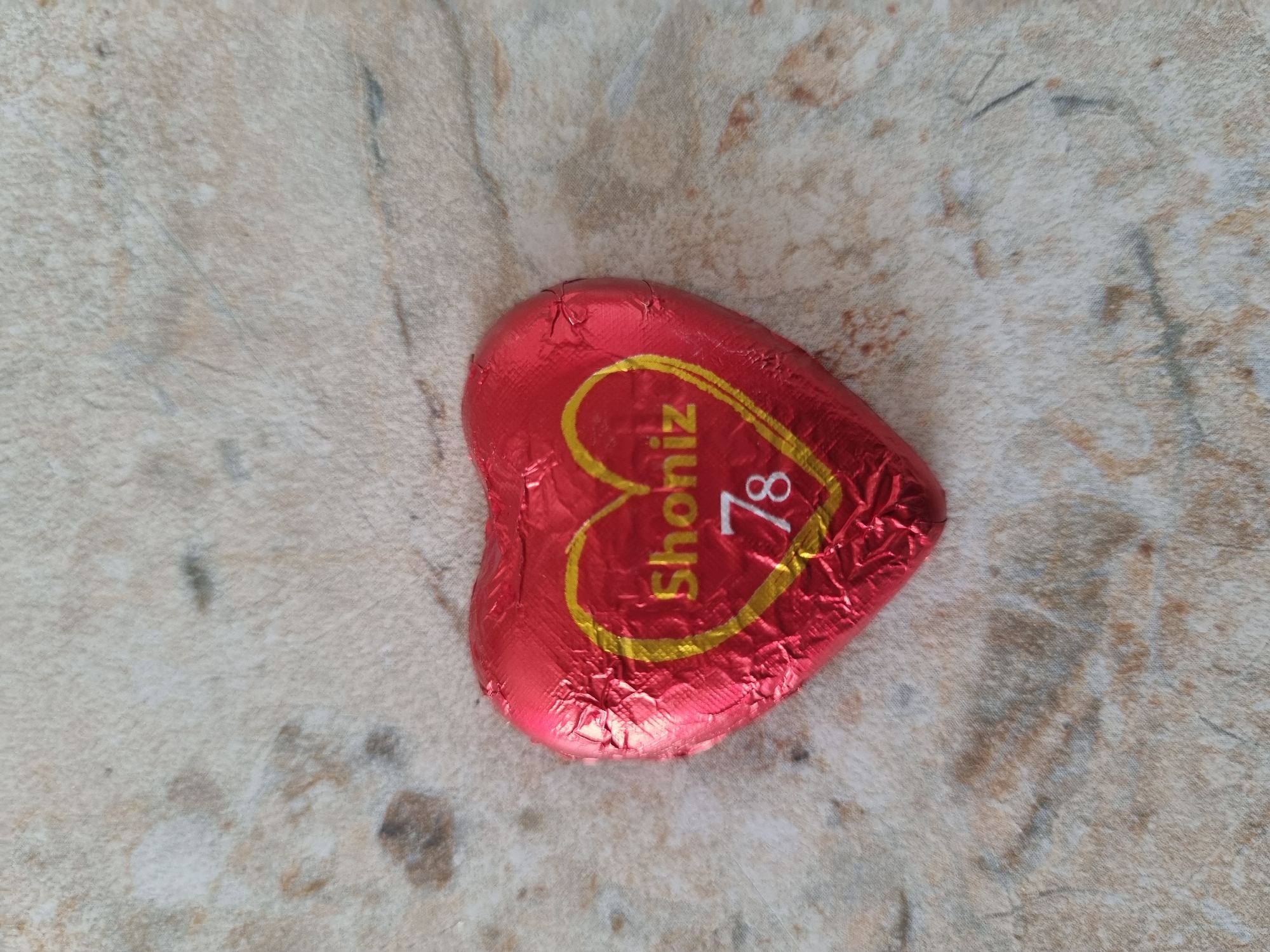 A heart-shaped chocolate candy, wrapped in crinkled red foil, rests on a mottled beige and brown surface that may be a countertop or floor. The foil has a gold-colored design that includes the text "Shoniz" and the number "78." The location is not identifiable as Sochi, Russia based on the visual information provided.