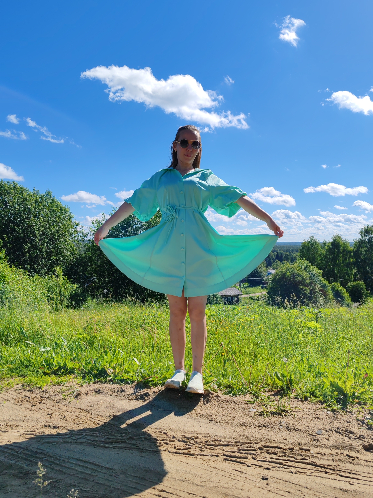 A young woman stands in a grassy field with a dirt path in the foreground. She is wearing a light blue, short-sleeved dress with a cinched waist and a floral embellishment on the side. The dress is buttoned down the front. She is also wearing sunglasses and white sneakers. The woman is holding the hem of her dress, flaring it out. Behind her, the landscape is lush and green, with trees and rolling hills visible in the distance. The sky is a vibrant blue with fluffy white clouds. The image appears to be taken outdoors in a rural setting, likely in or near the city of Chukhlem, Russia, given the overall aesthetic of the countryside. The light suggests it is daytime, likely during warmer months.