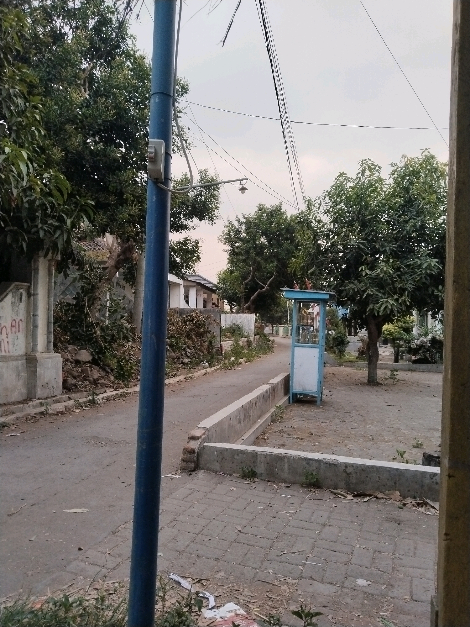 In Sangkanjaya, Indonesia, a street scene unfolds with a paved road curving to the left, flanked by a low concrete barrier. On the right, a small blue and white kiosk stands on a paved area, surrounded by trees and vegetation. A prominent blue pole is positioned in the foreground on the left, with a small electrical box attached to it. Beyond the kiosk, trees and houses are visible, with overhead power lines crisscrossing the sky. The overall ambiance is that of a quiet, residential street with a touch of local commerce.