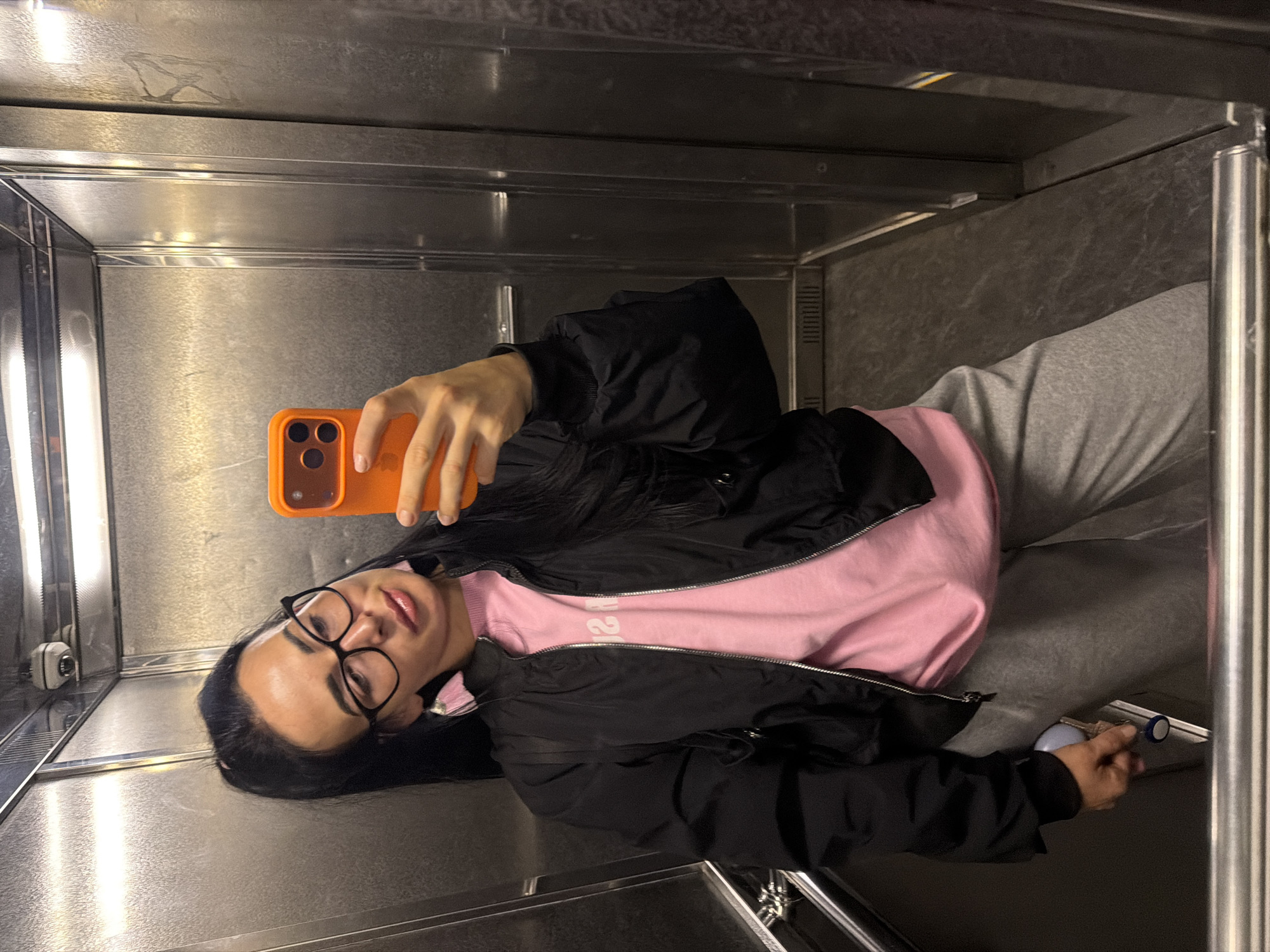 A woman stands in what appears to be a metal elevator, taking a selfie with an orange cell phone. She wears glasses, a pink top, a black jacket, and grey sweatpants. She holds a key in her other hand. The elevator is metallic and appears to be utilitarian. The location is Saint Petersburg, Russia.