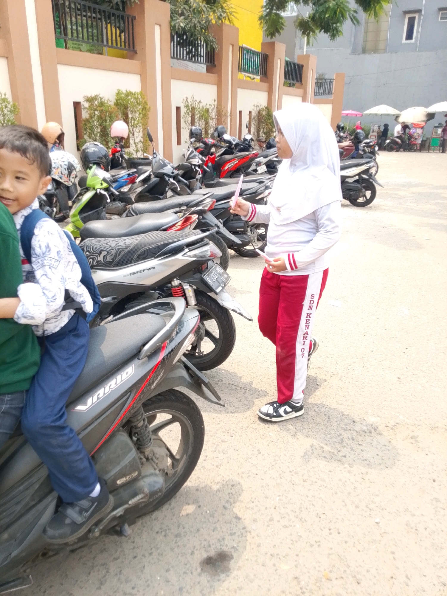 In Central Jakarta, Indonesia, a young boy wearing a white and blue patterned shirt, blue pants, and black shoes is sitting on a dark grey motorcycle. Next to him stands a girl in a white hijab and red and white striped pants, holding a small pink object. She is wearing black and white sneakers. In the background, many other motorcycles are parked, and a building with tan and brown accents is visible. Trees and foliage can be seen around the building. A few people are also visible in the distance, near some stalls with umbrellas. The scene appears to be an outdoor parking area on a sunny day.