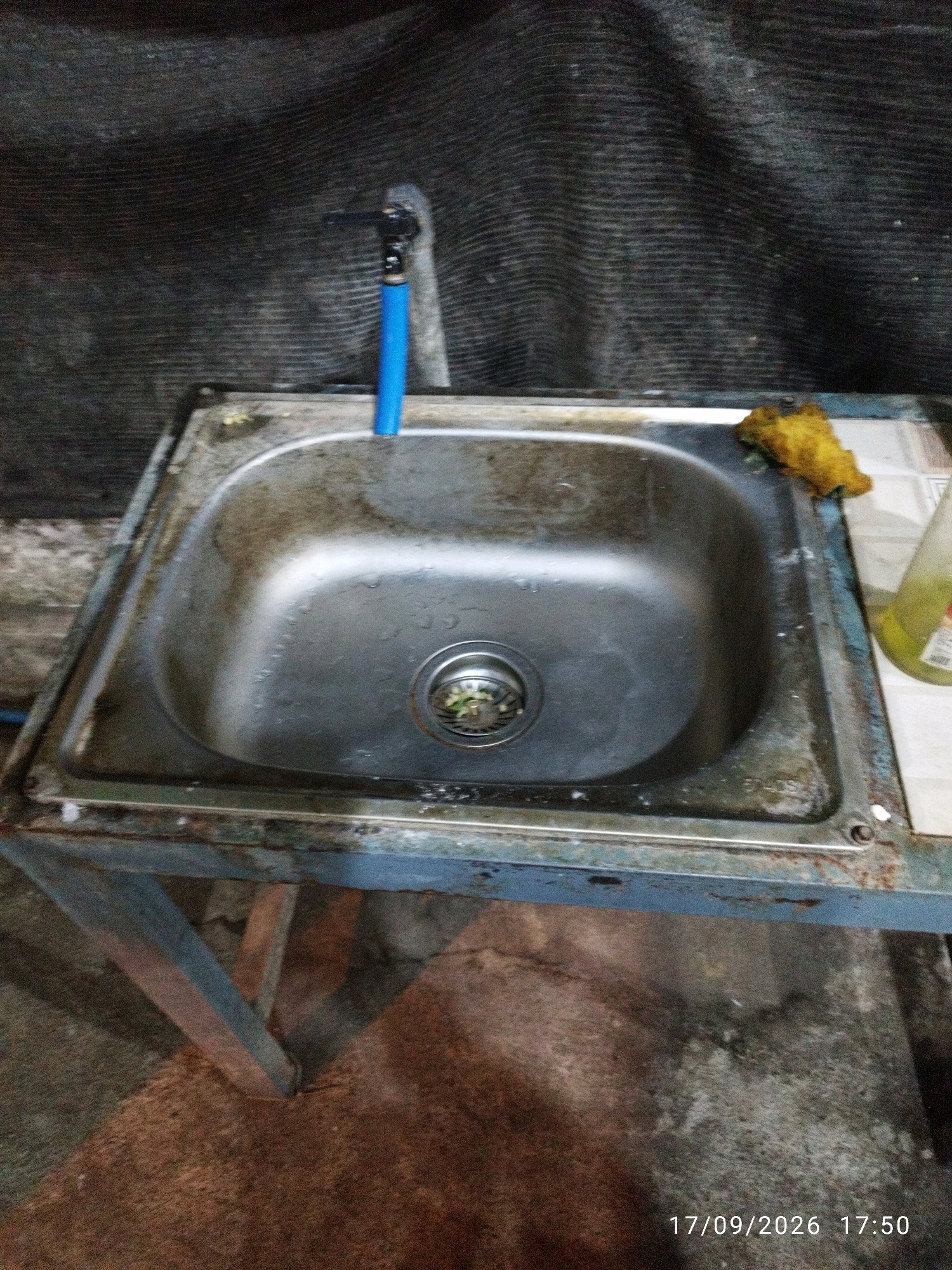 An outdoor sink area is depicted, likely in Mojokerto, Indonesia, based on the timestamp. The focus is on a stainless steel sink with a faucet attached to a blue pipe. The sink appears to be used, with some residue and water visible inside. A yellow sponge rests on the right edge of the sink. The sink is set into a metal frame which is supported by angled metal legs, suggesting it might be part of an outdoor preparation or washing station. The background consists of a dark, textured fabric or netting, possibly for shade. The ground beneath the sink is rough and appears to be concrete or dirt. A bottle of cleaning liquid is partially visible to the right of the sink. The overall scene suggests a functional, utilitarian space.