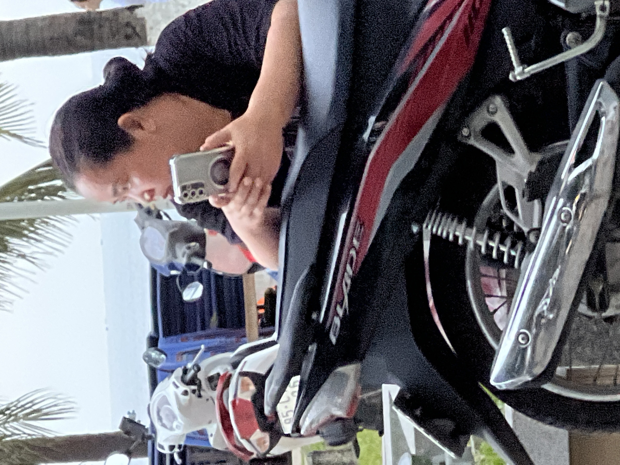 An Asian woman, with her hair pulled back in a bun, sits on a motorcycle. She is wearing a black t-shirt and is holding a smartphone in her hands, appearing to be looking at it. The motorcycle has a black seat and a red and black design on its body. Other motorcycles are visible in the background, along with palm trees and what appears to be the ocean or a large body of water. The scene suggests a public space in Nha Trang, Vietnam, likely a roadside or parking area near the coast.
