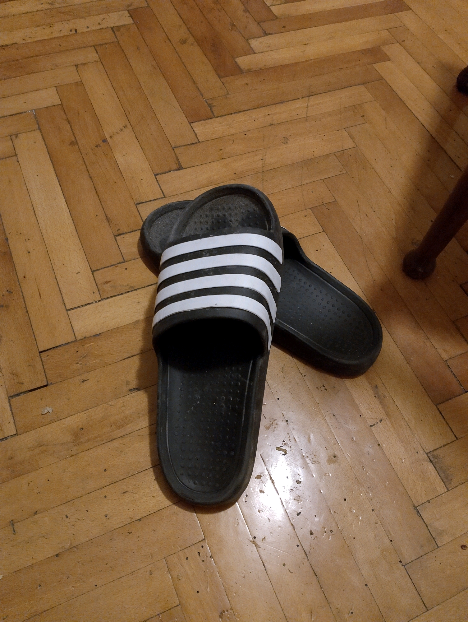 The image shows a pair of black Adidas slide sandals with white stripes resting on a herringbone patterned wooden floor. The floor appears to be aged and slightly worn, with some visible dust and debris. The sandals are placed close together, with one slightly overlapping the other. The lighting suggests an indoor setting, possibly a room in a home. There is no visible indication of the city of Herceg Novi, Montenegro. The scene is static, with no discernible actions or interactions taking place.