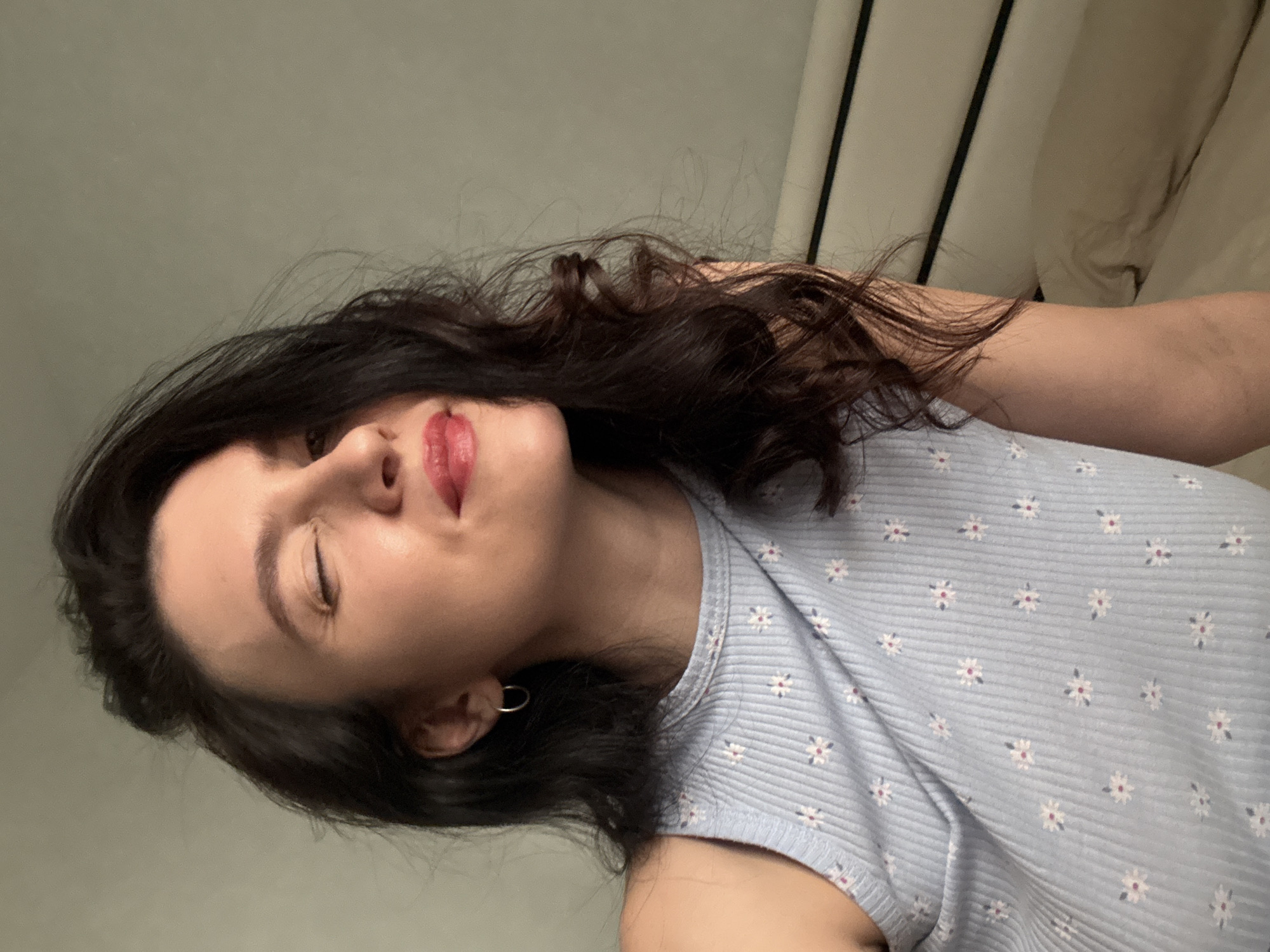 A woman with dark hair and fair skin is facing the camera with her head tilted to the left. Her eyes are closed, and she is wearing red lipstick. She is wearing a light blue shirt with a floral pattern. A silver hoop earring is visible in her left ear. The background consists of a light gray wall and a white headboard. There is no indication of the location being in Kazan, Russia.