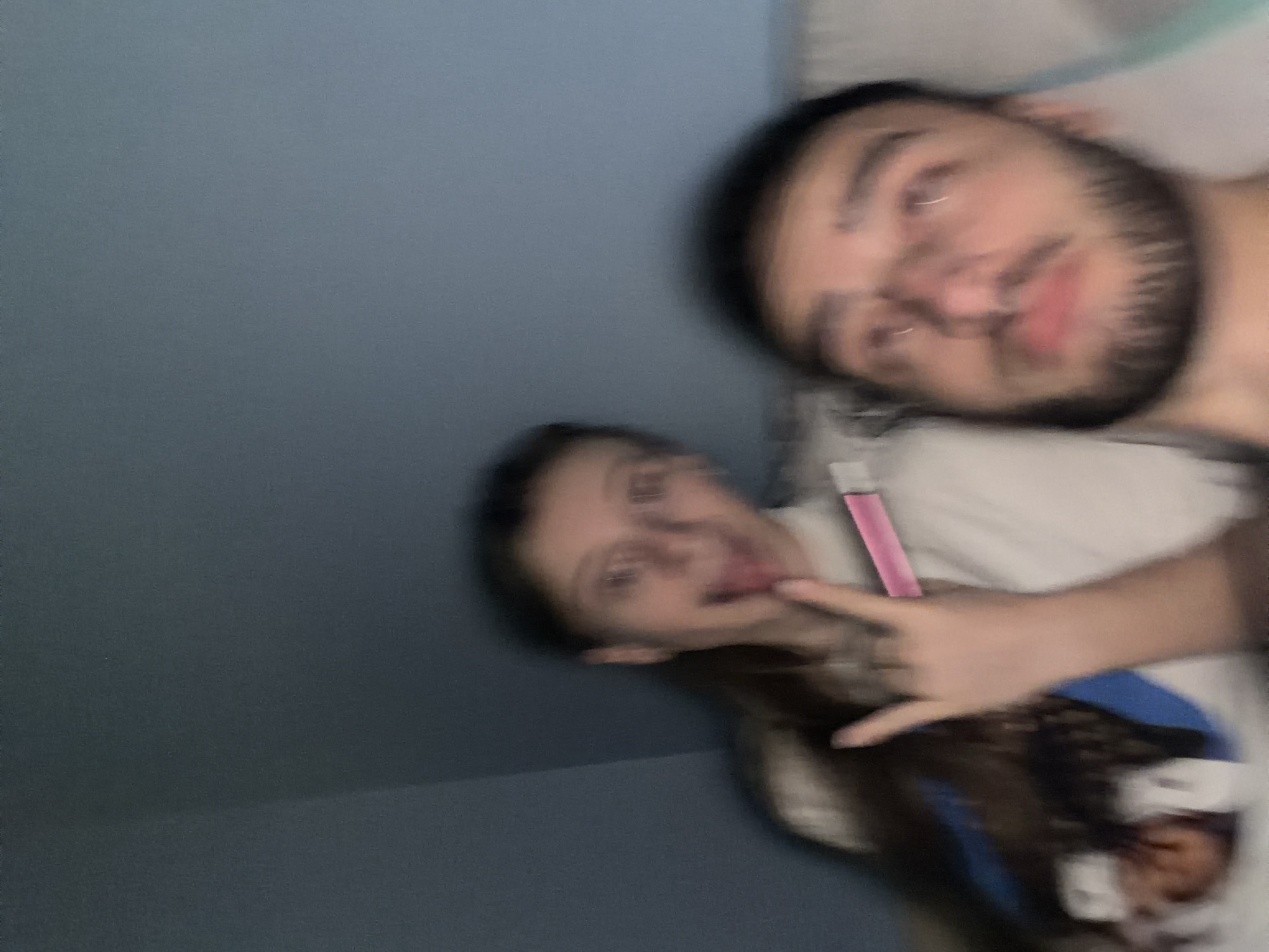 Two individuals, a man and a woman, are captured in a blurry, close-up shot. The man has a beard and facial piercings. The woman is sticking her tongue out and making a hand gesture. She is holding a pink object. They are positioned against a blue wall and an area that appears to be bedding. Location: Undetermined, not possible to establish from the image. There is no evidence to suggest the location is Plovdiv, Bulgaria.