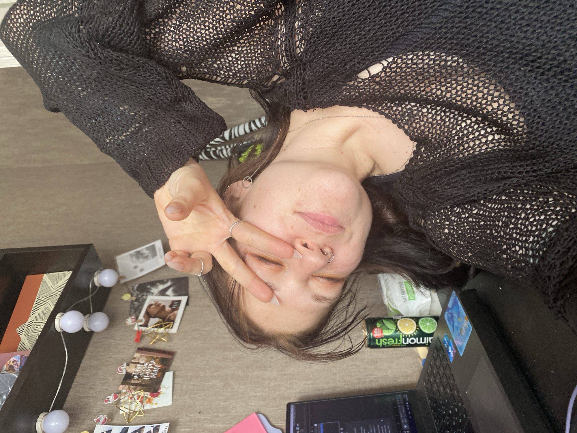 A woman is lying on a desk or floor, facing the camera. She is wearing a black, loosely knitted top and has brown hair. Her left hand is raised near her face, with her fingers partially obscuring her eye. She has a nose ring and rings on her fingers. A laptop is visible to her right, with a screen displaying what appears to be code. Next to the laptop, there is a package with "Limonfresh" written on it. To her left, a collection of photos is arranged near a decorative shelf with string lights. Based on the brand name on the package ("Limonfresh"), and the assumption the name represents the location of the packaging company, the location of this photo is likely Chaikovsky, Russia.