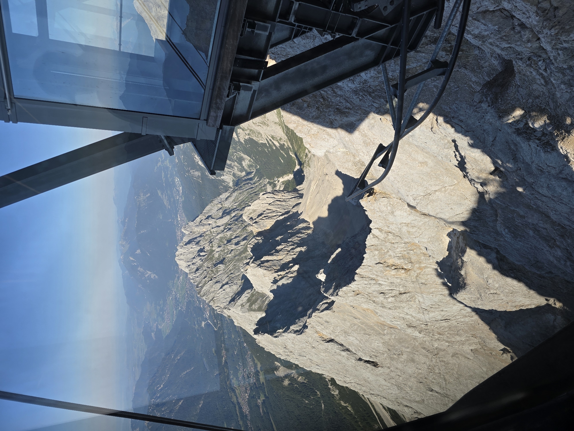 The image depicts a panoramic view from a high vantage point, likely a cable car or observation platform, overlooking a mountainous region. In the foreground, the metallic structure of the observation point, including beams, cables, and glass panes, is visible, framing the landscape. The mountains dominate the scene, with jagged, rocky peaks casting long shadows due to the low angle of the sun. The texture of the rock faces is rugged and varied. In the middle ground, a valley stretches out, dotted with houses and green vegetation, indicating a settlement nestled within the mountains. The distant background shows more layers of mountains fading into a hazy horizon under a clear blue sky. The overall impression is one of immense natural scale and a high altitude perspective. The context provided indicates this view is from Garmisch-Partenkirchen, Germany, suggesting a likely location at a prominent mountain peak accessible by cable car, such as the Zugspitze.