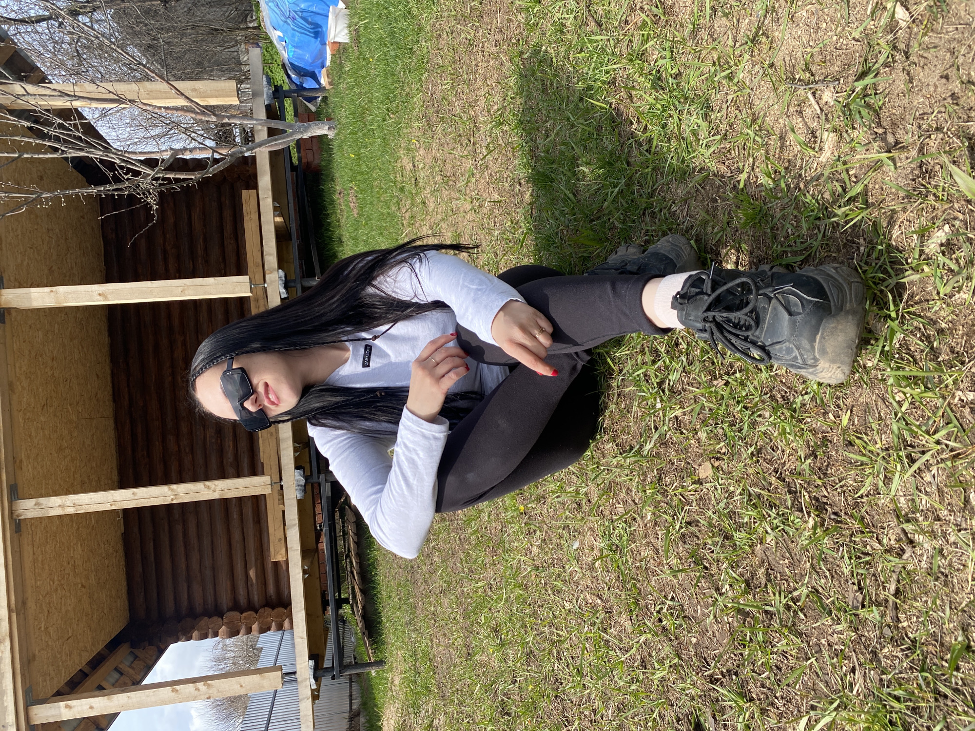 A woman sits on the ground in Chaikovsky, Russia. She has long, dark hair with thin braids woven in, and she is wearing sunglasses, a white long-sleeved shirt, black leggings, and black boots. She is seated in a grassy area with patches of dirt. Behind her is a partially constructed wooden structure, likely a shed or small house, and a bare tree. A blue tarp is visible in the background. A metal fence runs along the bottom edge of the frame.