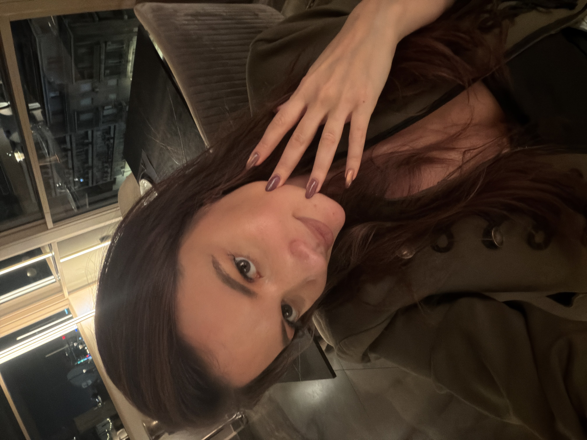 The image features a woman posing indoors at night, with her head slightly tilted and direct eye contact towards the camera. Her long, dark brown hair cascades around her shoulders. She is wearing a dark, possibly olive green, jacket or top with button details. Her right hand, adorned with metallic reddish-brown/purple nail polish and hints of gold/copper on some nails, is gently resting on her hair near her forehead.
The indoor setting is dimly lit, suggesting an evening or nighttime ambiance. Behind her, a dark, textured, upholstered surface, likely a sofa or chair, is visible. Through a large window to her left, an urban night scene is reflected, showing illuminated building facades and various street or window lights from other structures, consistent with a city environment like Athens, Greece. Rectangular interior light fixtures are also reflected in the glass. The overall scene conveys a relaxed moment in what appears to be an upscale urban establishment. No visible text is present in the image.