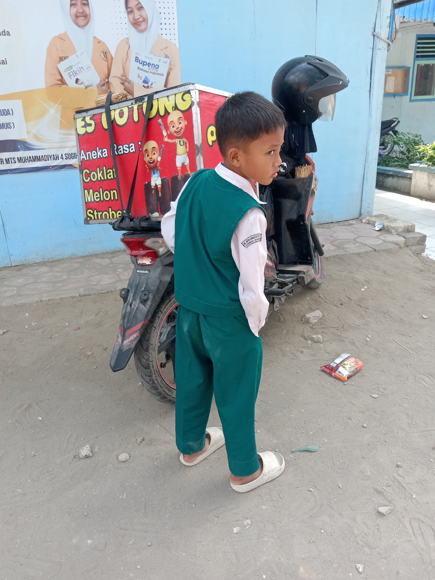 A young boy, appearing to be of Indonesian ethnicity, stands next to a red ice cream cart attached to a black motorcycle. He is wearing a white collared shirt and a green vest, along with green pants and white sandals. His hair is short and dark. The ice cream cart displays various flavors, including "Aneka Rasa," "Coklat," "Melon," and "Stroberi," and features cartoon characters. A black helmet rests on the back of the motorcycle. The ground is sandy with scattered stones. In the background, a light blue building with windows is visible, and further in the distance, there is greenery and a parked motorcycle. A discarded packet of snacks lies on the ground in front of the boy. The scene is set in Sugihwaras, Indonesia, indicated by the text on the boy's shirt which reads "MTS MUHAMMADIYAH 4 SUGIHWARAS."