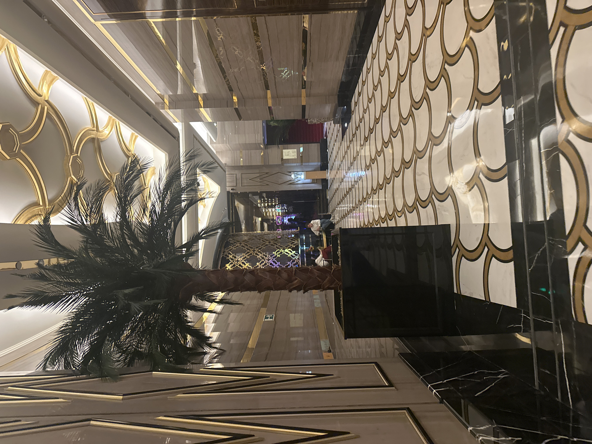 The image depicts a luxurious indoor hallway with a prominent palm tree in a black planter. The hallway features a marble floor with an intricate white and gold scalloped pattern. The walls are adorned with marble panels and vertical gold accents. The ceiling is elaborately decorated with gold trim and recessed lighting. In the background, several people are visible, some seated and others standing. One person is seated near the palm tree, and two individuals are in the distance towards the right. A red carpet leads up a set of stairs further down the hallway. The overall impression is one of opulence and grandeur, consistent with a high-end hotel or casino. The location is identified as Estosadok, Russia.