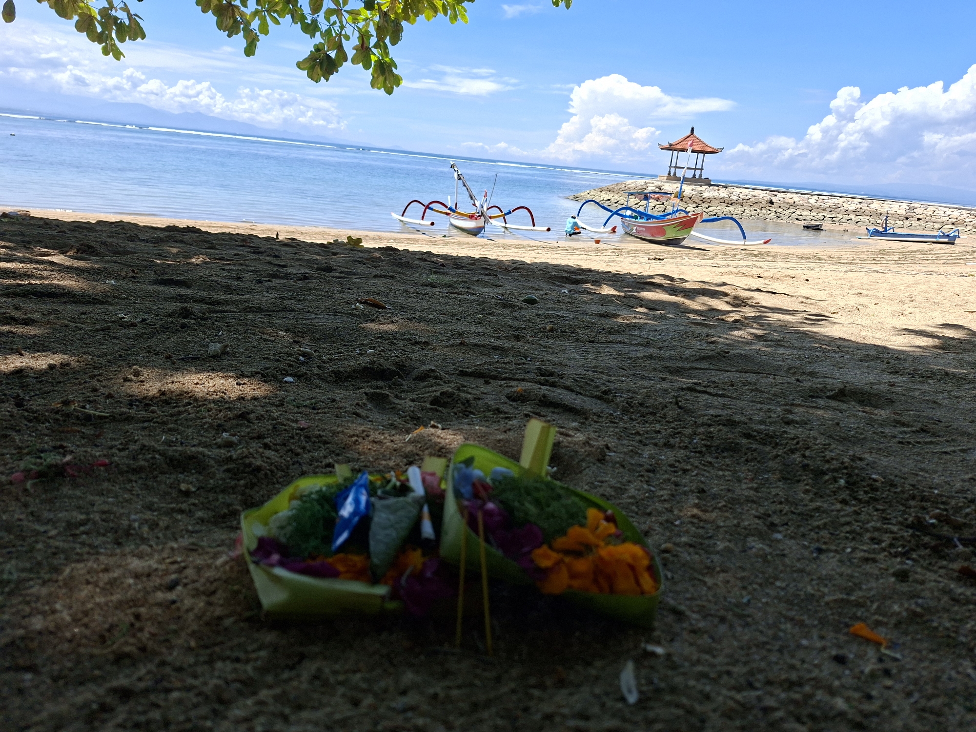 This wide-angle shot captures a serene beach scene in Denpasar, Indonesia, under a bright, clear blue sky with scattered white clouds. The foreground features dark, damp sand, partially shaded by green tree leaves hanging into the frame from above. Two traditional Balinese offerings, known as Canang Sari, are placed on the sand in the immediate foreground. These offerings are small woven palm-leaf baskets filled with colorful flowers and other items, appearing slightly out of focus.
Further back, the sandy beach meets the calm, turquoise ocean. Several traditional Balinese fishing boats, or jukungs, with their distinctive outriggers and vibrant colors, are pulled up onto the sand near the water's edge. One prominent jukung in the mid-ground has red and white outriggers and a mast. Another colorful jukung is seen to the right, closer to a stone breakwater that extends into the sea. At the end of the breakwater stands a small, open-sided gazebo or balé with a red-tiled roof.
Beyond the breakwater, the ocean stretches to the horizon, where faint outlines of distant land or mountains are visible. The bright sunlight casts distinct shadows on the sand, indicating a clear, sunny day, likely late morning or early afternoon. The scene evokes a peaceful, tropical atmosphere characteristic of Bali. No people are visible, and there is no discernible text within the image.