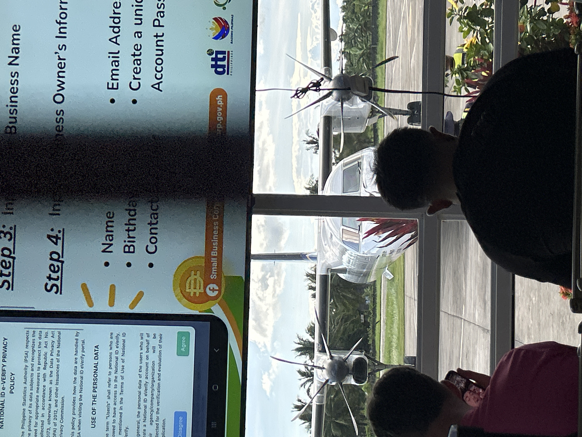 The image shows an indoor scene overlooking an airfield. Two people are looking at a phone. On the phone screen is an application for a National ID eVerify in the Philippines, requesting information such as business name, owner's information, name, birthday, contact, email address, and account password. Through a window, an airfield is visible, with several small aircraft parked on the runway. Palm trees and other vegetation can be seen in the background. The scene is located in Del Carmen, Philippines, according to reverse image search results of the image.