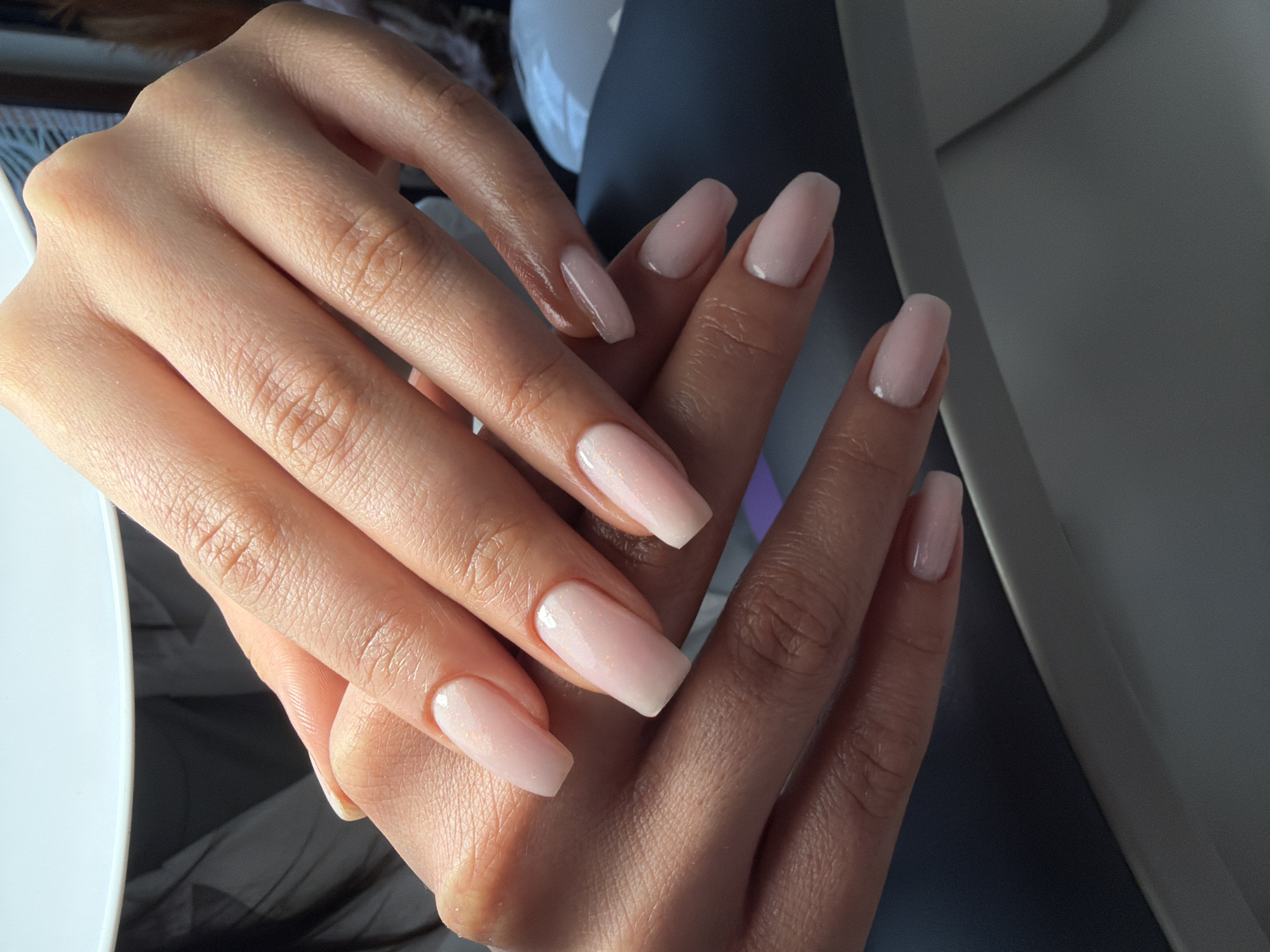 The image shows two hands with light pink nail polish, resting on what appears to be a light-colored surface. The nails are square-shaped and medium length. The skin tone is fair. The background is blurred, but suggests an indoor setting. There is no indication of the location being Burgas, Bulgaria.