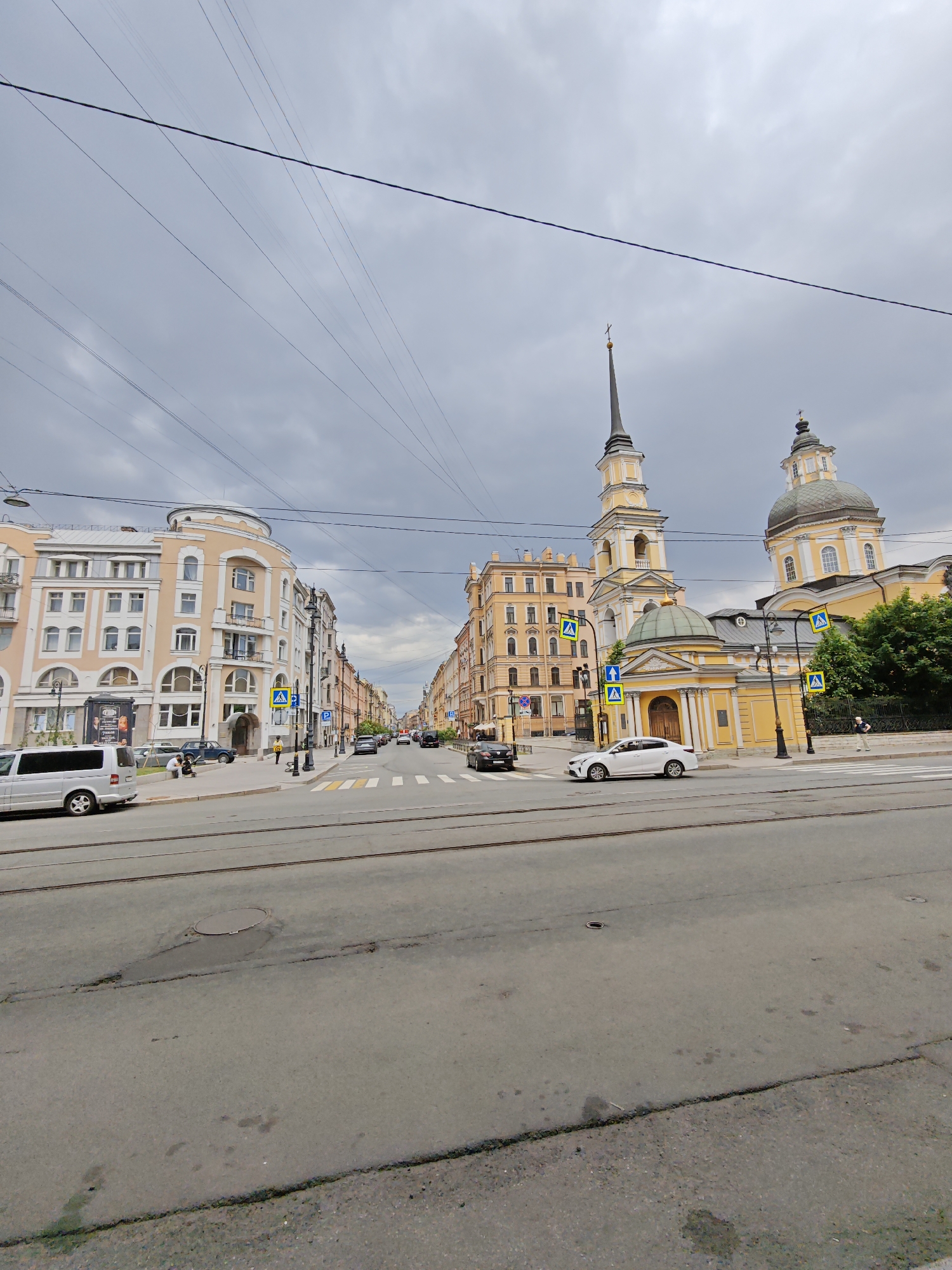 The image depicts a street scene in Saint Petersburg, Russia, on an overcast day. In the foreground, a wide asphalt road with tram tracks runs horizontally across the frame. The road shows some signs of wear, with visible cracks and a manhole cover.
To the left, a light-colored multi-story building with rounded architectural features and arched windows dominates the side of the street. A white minivan is parked to the left of this building, and a few people are seated on a bench on the sidewalk. Further down the street, more buildings with varying architectural styles and colors line the road. Pedestrian crossings with yellow "zebra" markings and blue directional signs are visible.
On the right side of the street, a prominent yellow church with a tall bell tower and a dome is visible. The church has a distinctive architectural style, with arched windows and decorative elements. A white car is driving past the church on the road. Trees and a black metal fence border the church property.
Overhead, numerous power lines crisscross the sky, creating a complex network against the gray, cloudy backdrop. The overall atmosphere is that of a typical urban street in Saint Petersburg, with a mix of historical and modern architecture.