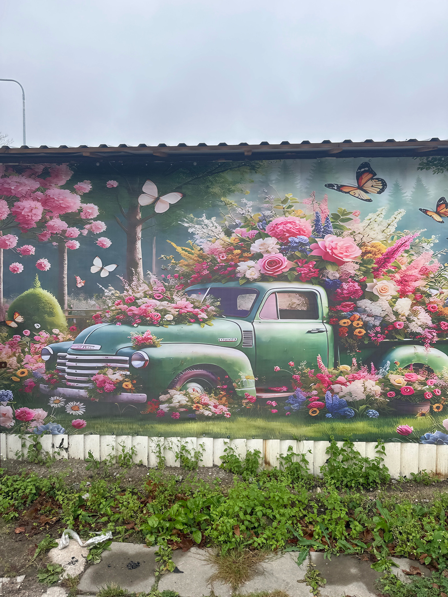 The image shows a vibrant mural painted on a wall. The mural depicts a vintage green pickup truck overflowing with a lush arrangement of colorful flowers, primarily pinks and purples, with some yellow and white accents. Several butterflies are depicted in flight around the truck and among the flowers. The background of the mural features trees and a soft, hazy sky. In front of the mural, there is a low, white picket-style fence, and beyond that, overgrown green weeds and some paving stones. The scene appears to be outdoors, likely on a cloudy day, given the overcast sky in the mural. The mural is located in Vilnius, Lithuania.