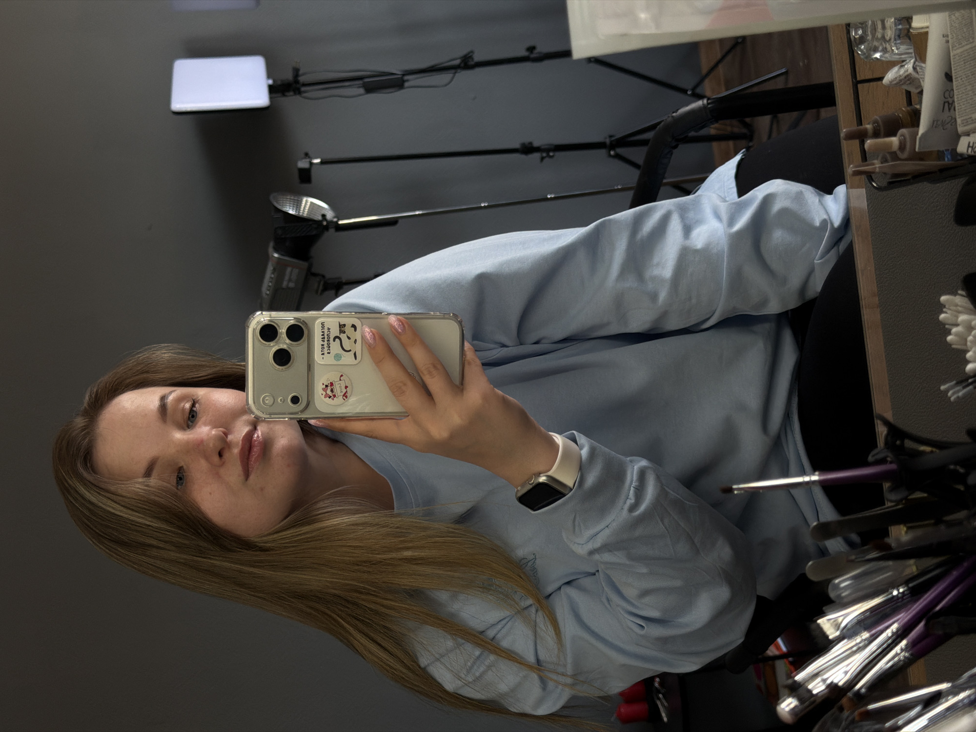 A fair-skinned young woman with long, light brown hair is taking a selfie with a cell phone in a clear case with stickers on it. She is wearing a long-sleeved, light blue shirt and an Apple Watch with a white band. The background includes professional lighting equipment, including a square LED panel and a camera flash mounted on tripods. To the right, a partially visible makeup station reveals cosmetic products and brushes. The scene appears to be indoors, possibly in a professional makeup or photography studio. The specific location is Chaikovsky, Russia.