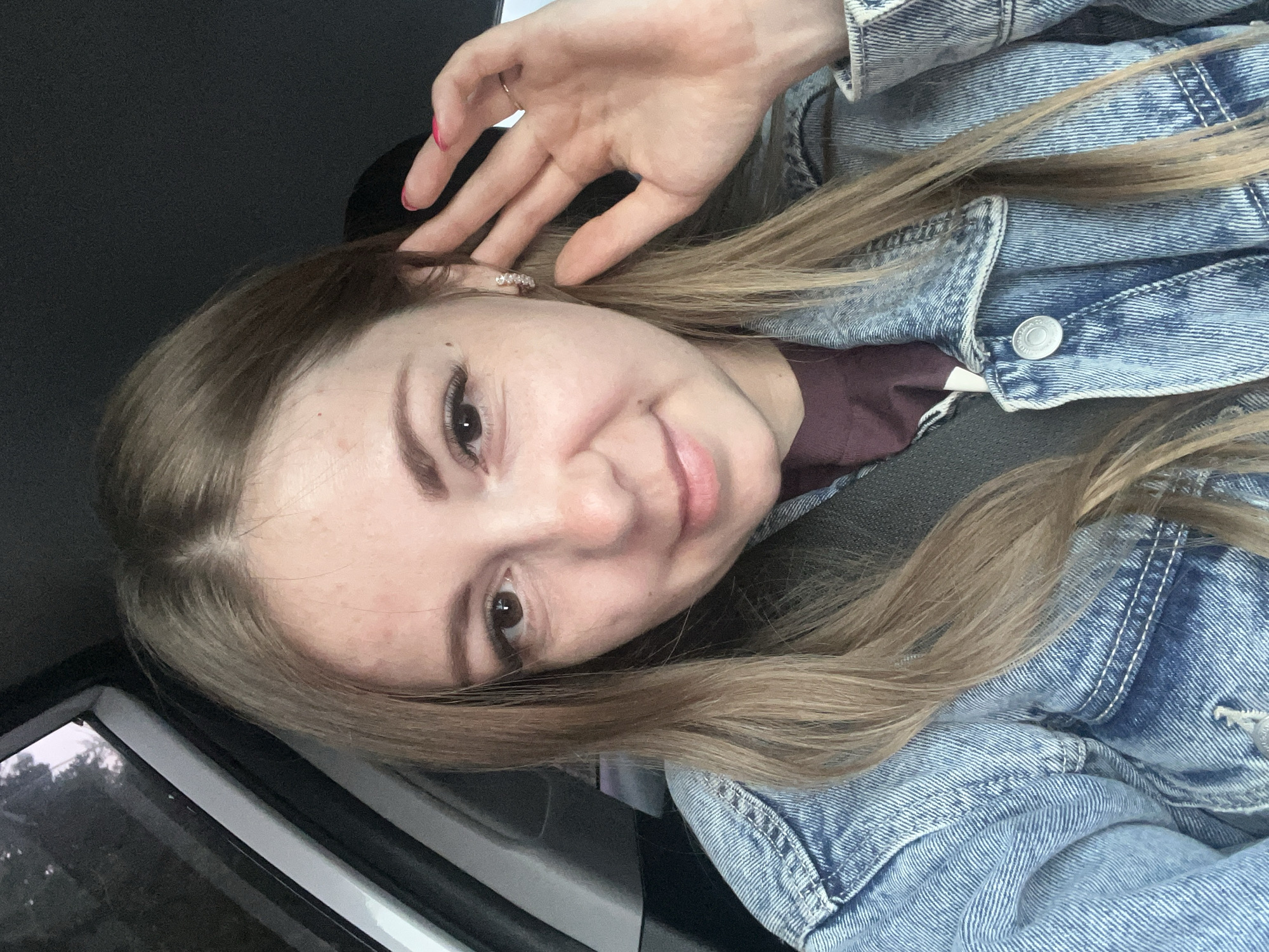 The image shows a close-up, selfie-style photograph of a young woman with long blonde hair, wearing a denim jacket over a burgundy shirt. She is in a car, as indicated by the seatbelt visible across her chest and the car window in the background. Her right hand is raised to her ear, and she is looking towards the camera with a slight smile. The lighting suggests it is daytime, possibly late afternoon or early evening. The background is dark and out of focus, with a hint of trees visible through the car window. There are no specific details that indicate the location in Allaki, Russia.